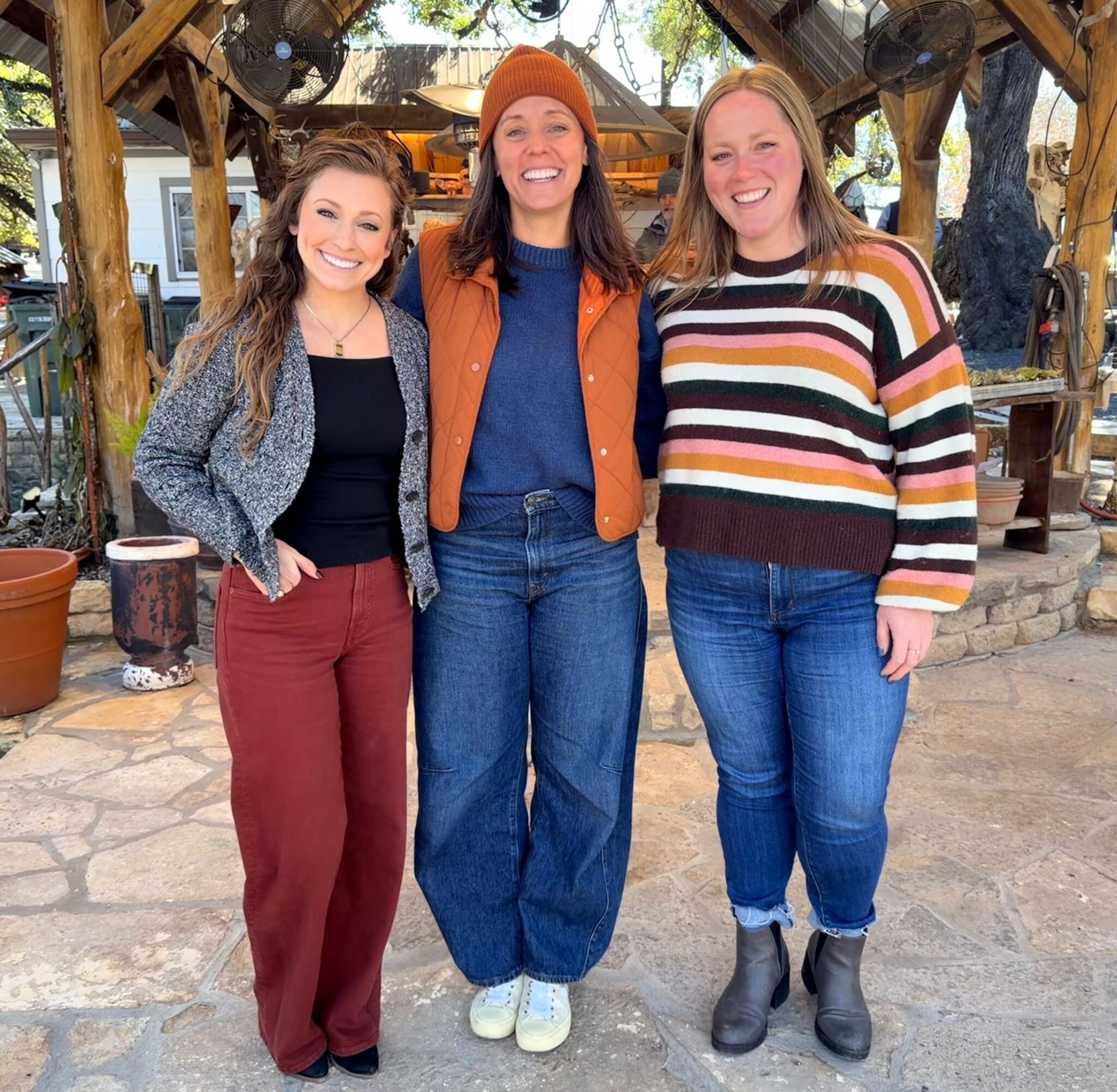 Three women standing outdoors, smiling, with wooden structures and plants in the background.