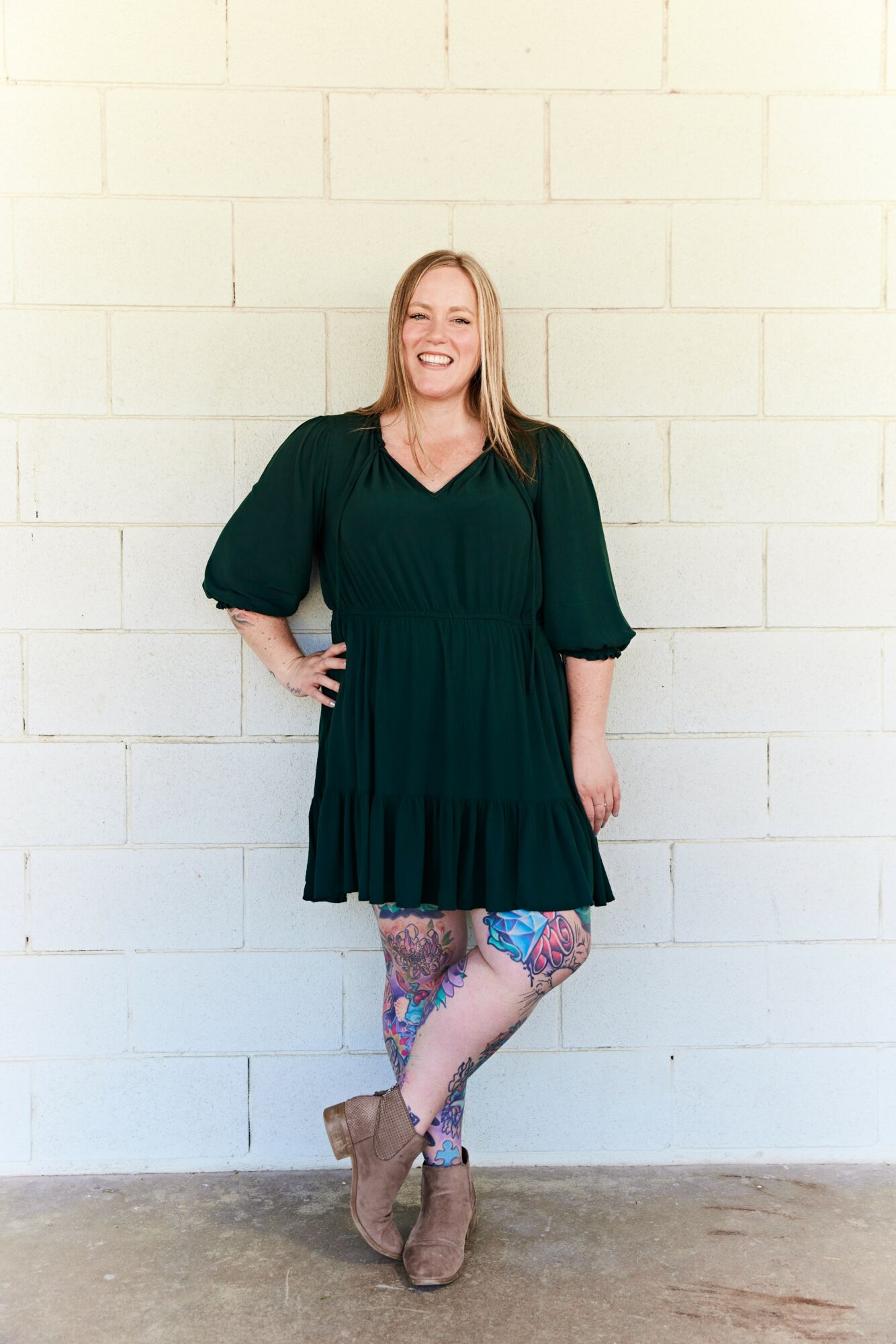 Woman with long blonde hair smiling, wearing a dark green dress and patterned tights, standing against a white brick wall.
