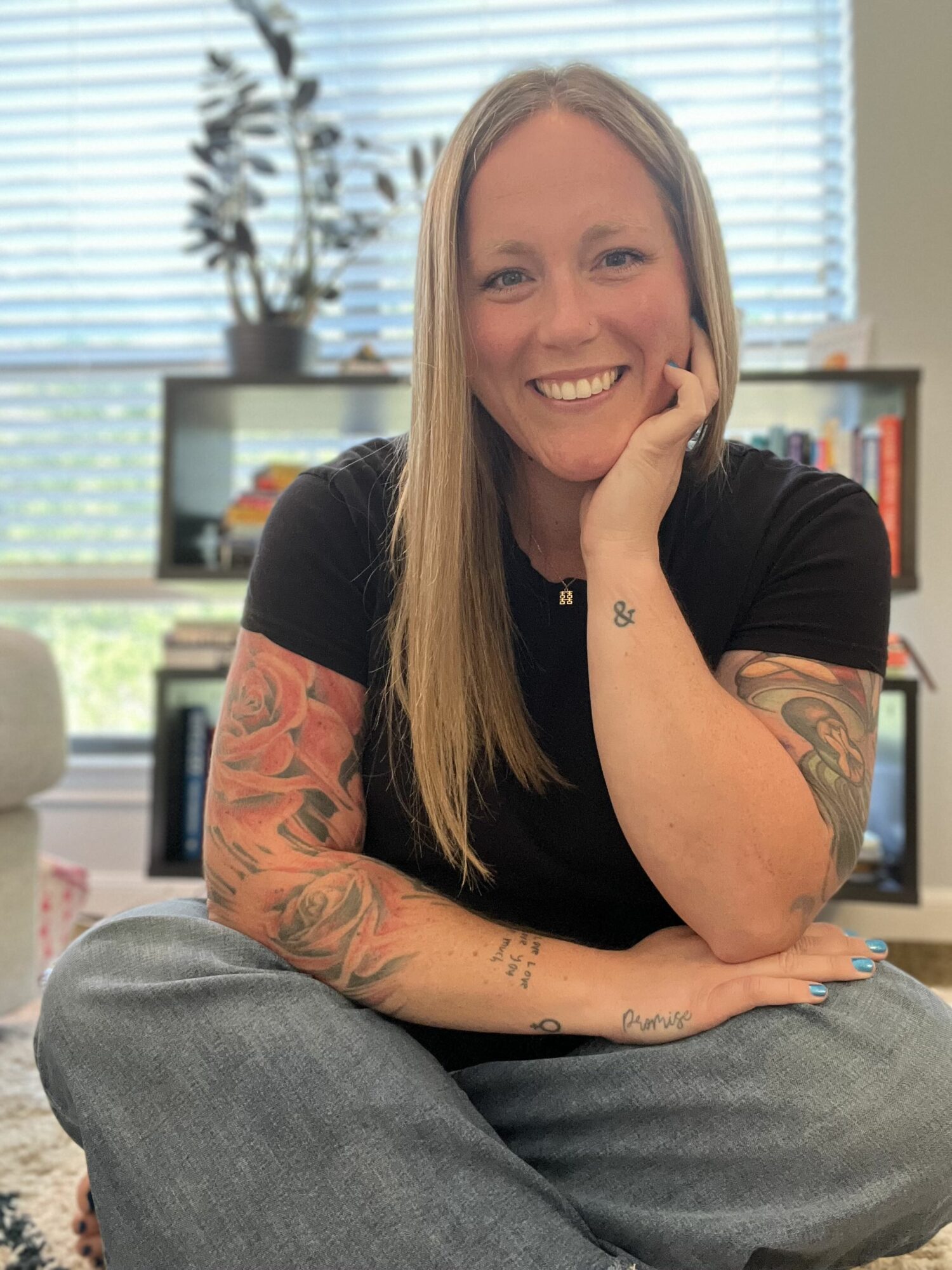 Smiling woman with long hair, tattoos on arms, sitting cross-legged indoors with bookshelves and window blinds behind her.