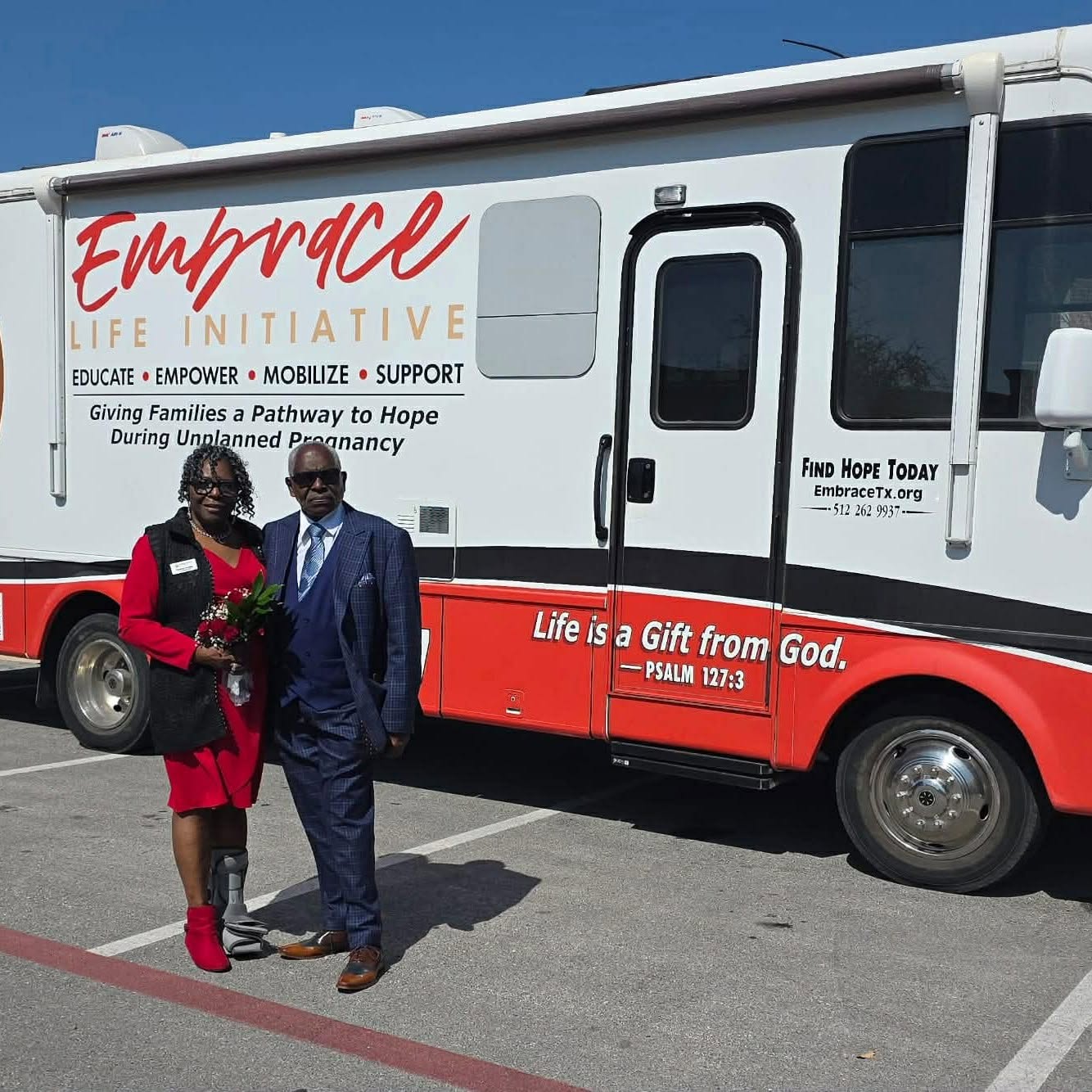 Two people stand in front of a mobile outreach vehicle with Embrace Life Initiative branding, on a parking lot, sunny day.