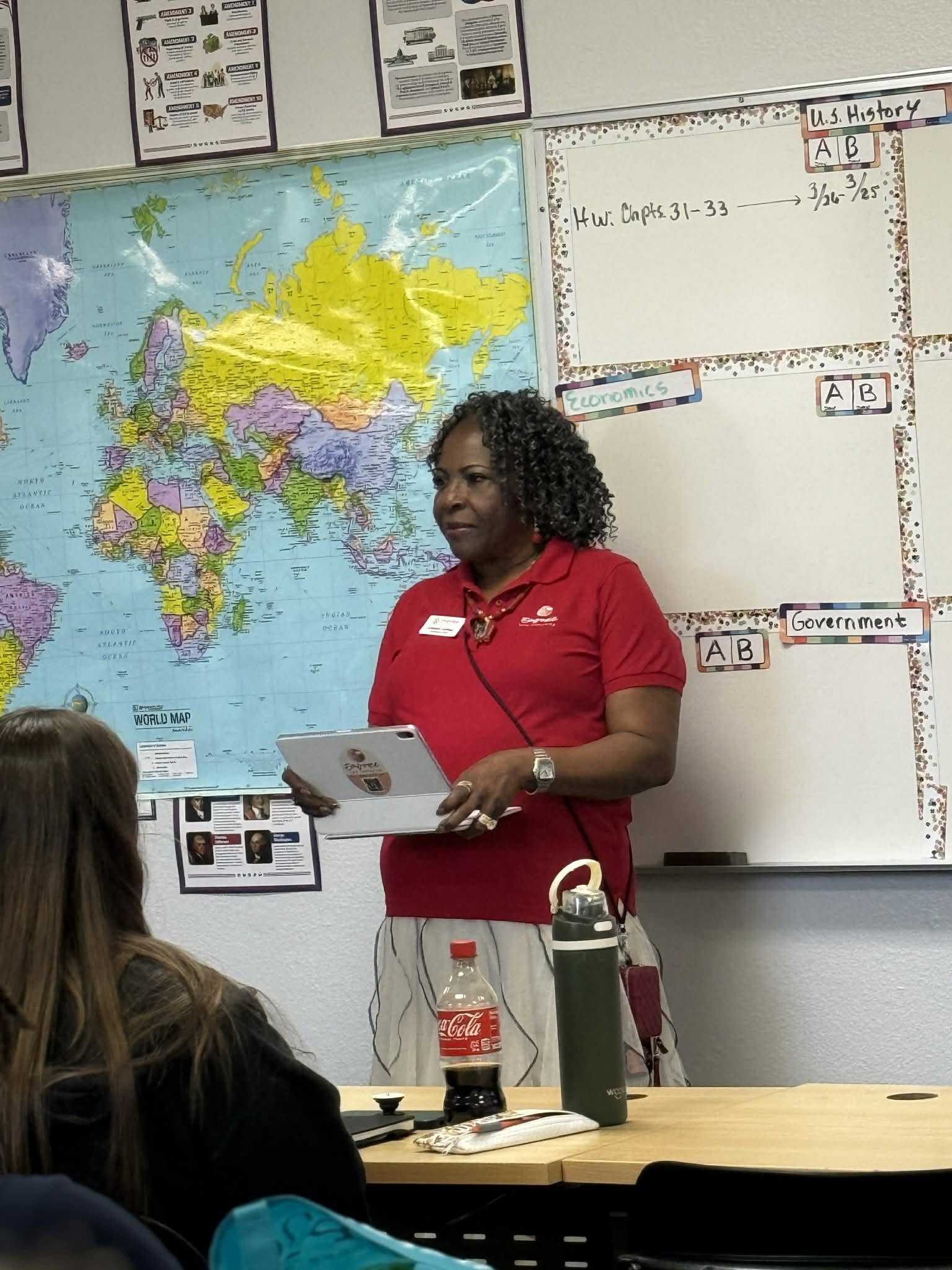 A woman with curly hair in a red shirt stands in front of a world map and whiteboard in a classroom.