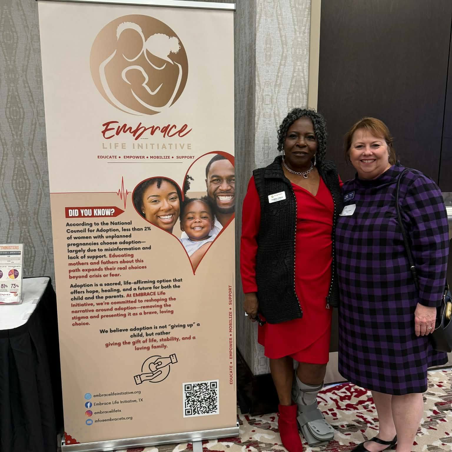 Two women stand together indoors, smiling, next to a vertical banner with a logo of a mother and child, and text about an initiative.