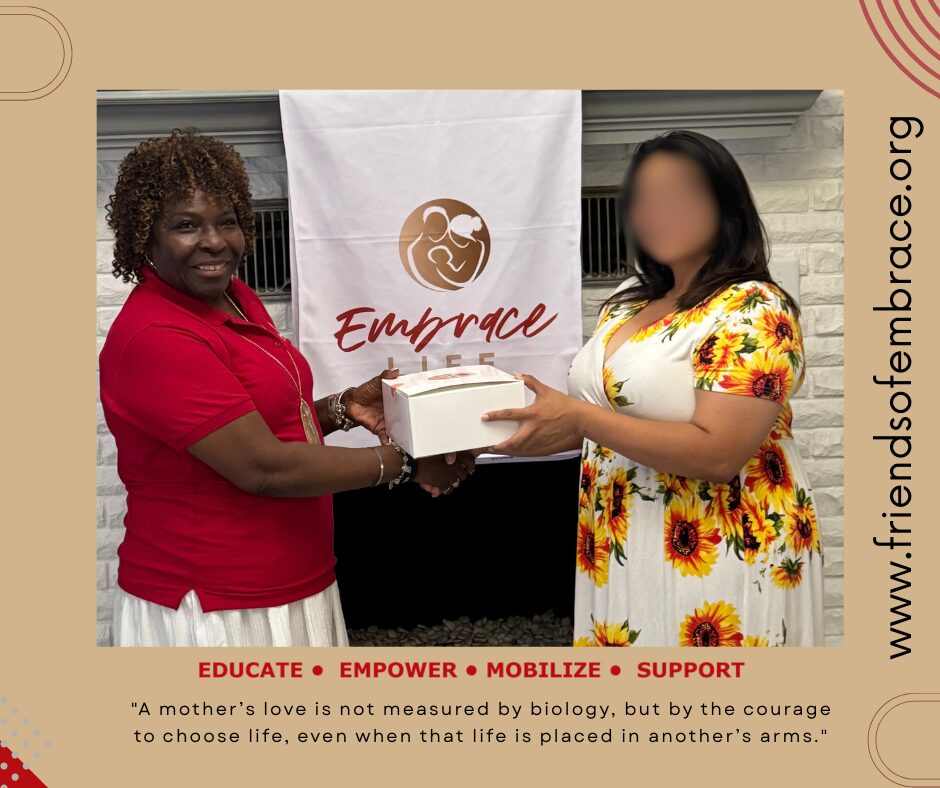 Two women exchanging a box in front of a banner with the word 'Embrace' and a logo, smiling. One woman wears a red shirt, the other a floral dress.