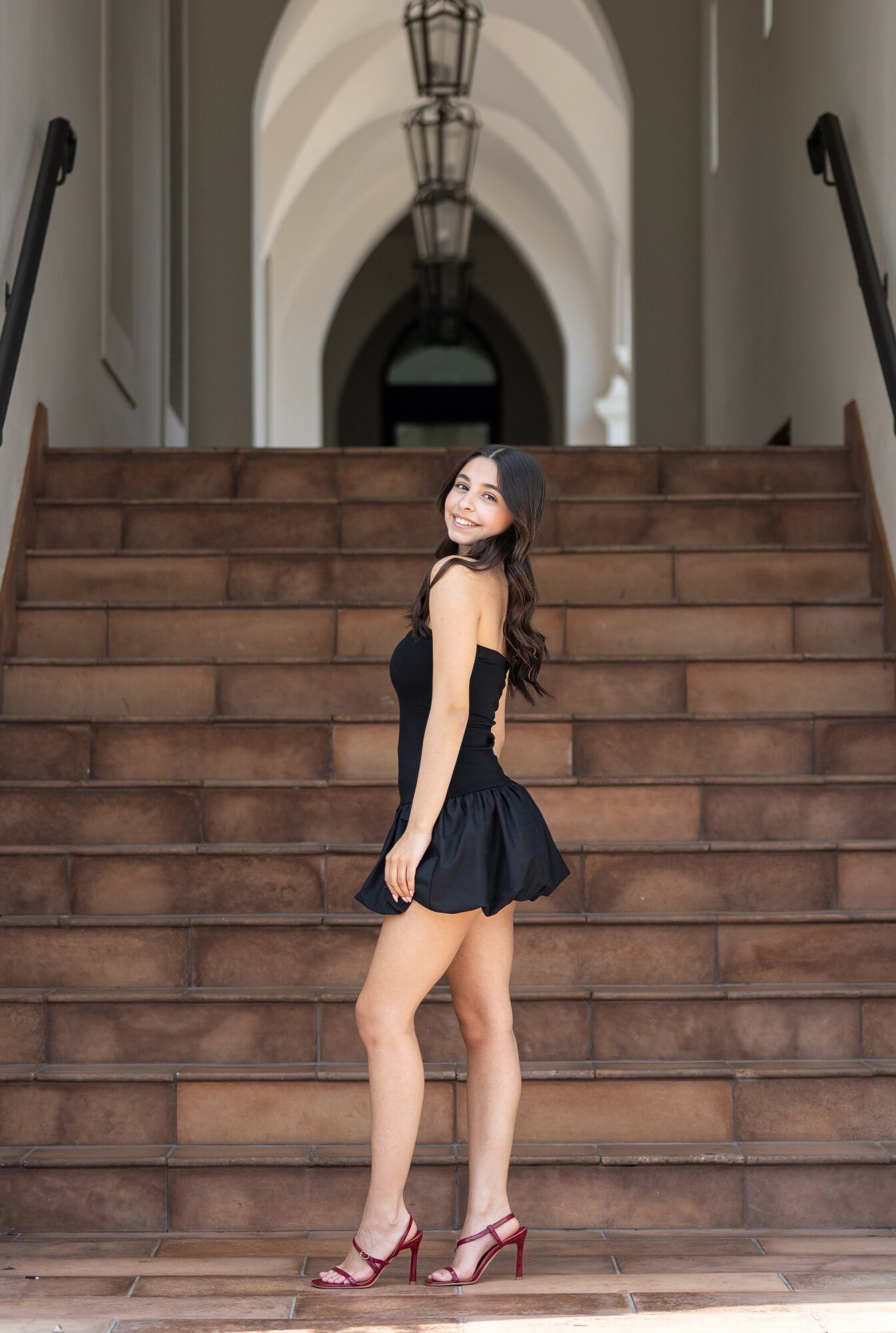 Young woman in black dress and red heels standing on stairs, smiling, in an indoor hallway with arched ceiling and hanging lights.