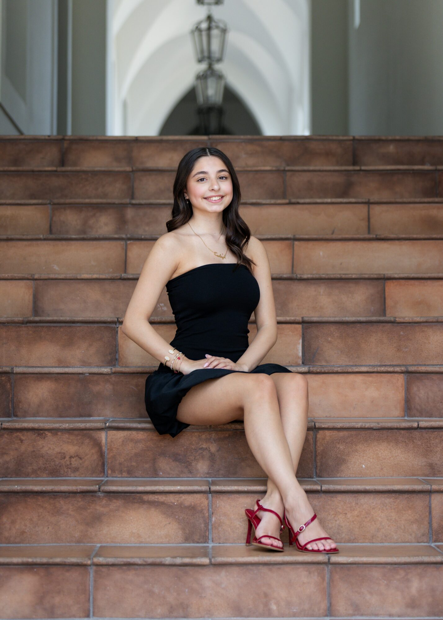 Young woman in a black strapless dress sitting on stairs, smiling, with a building and archway in background.