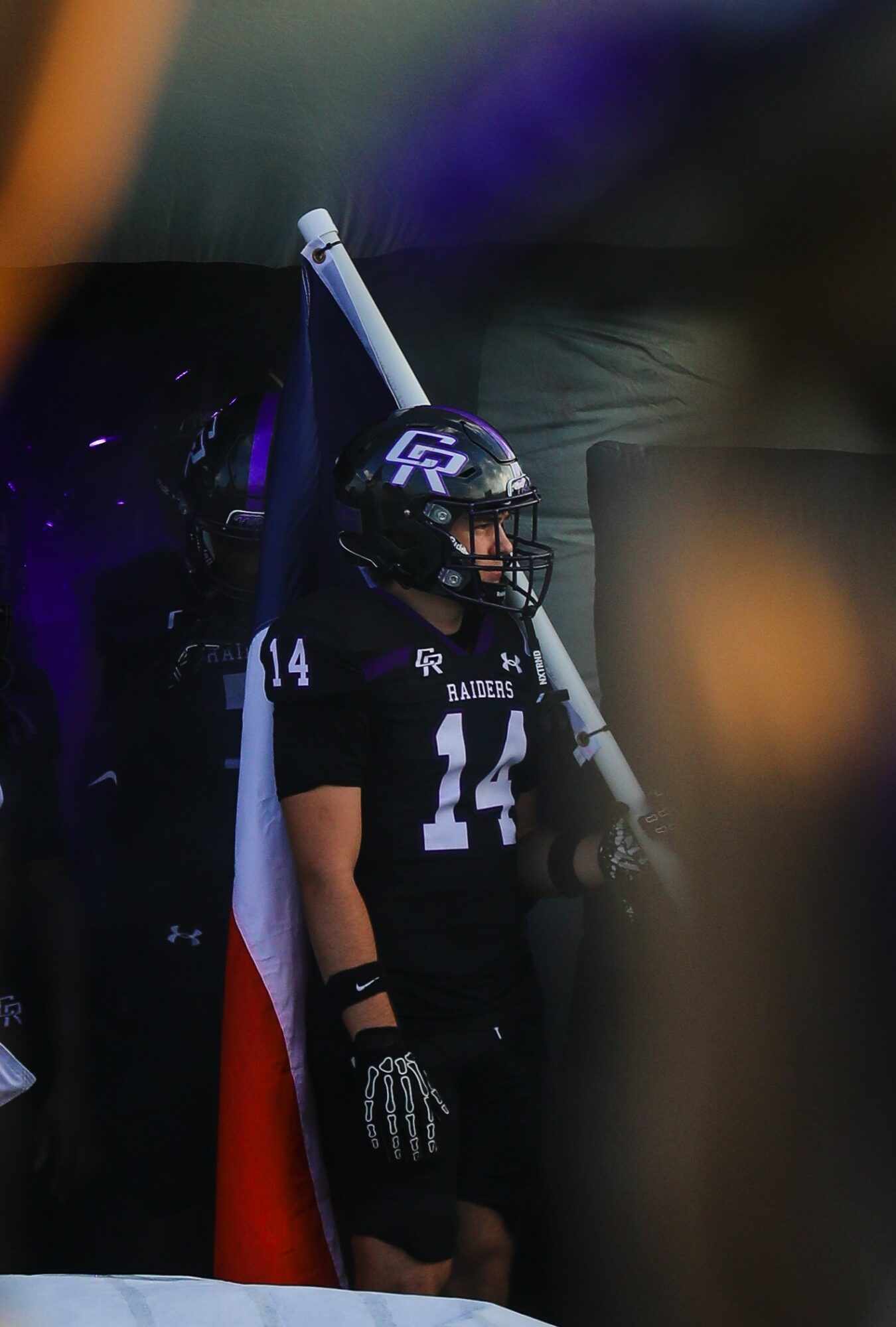 Football player in black uniform with number 14, wearing helmet, standing with flag behind, dark background.
