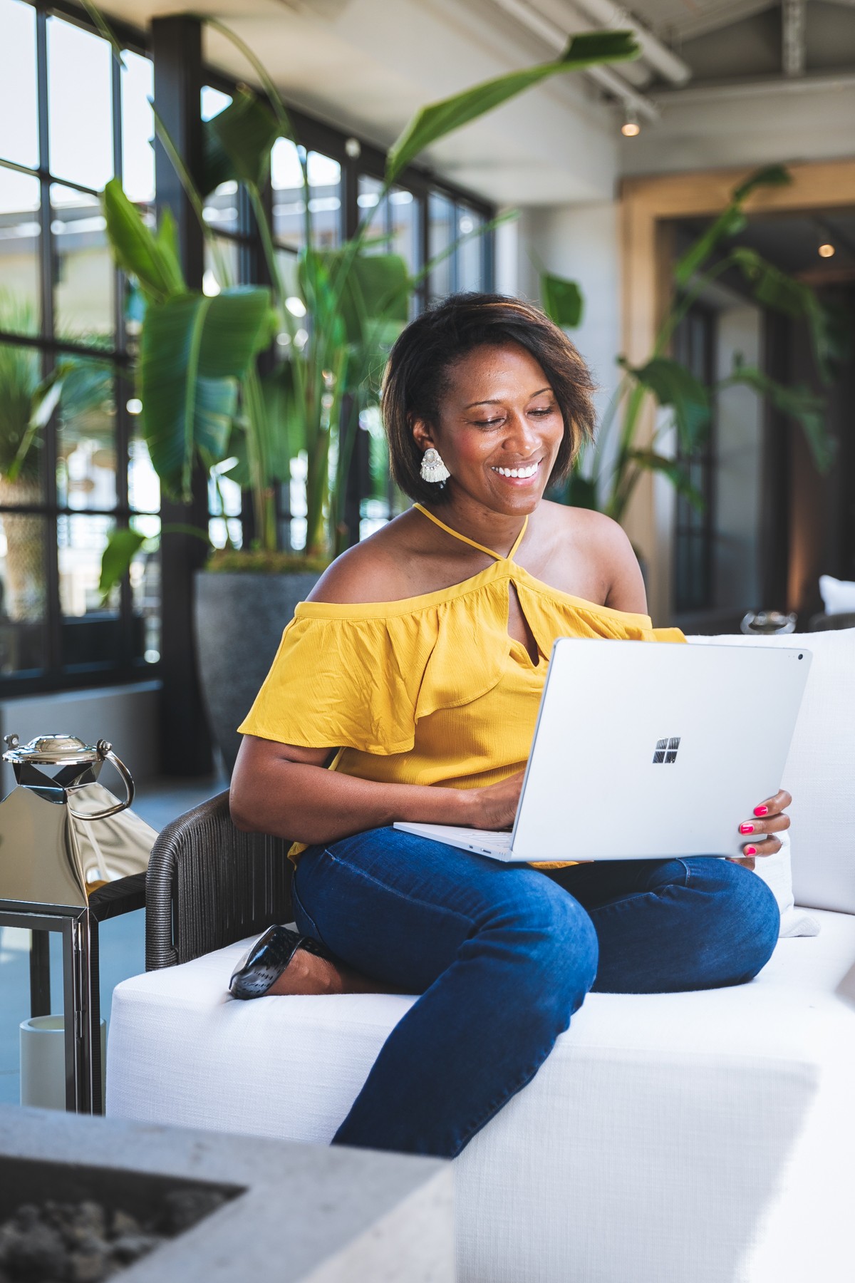 Woman sitting on a white sofa using a laptop in a modern indoor space with large plants and windows.