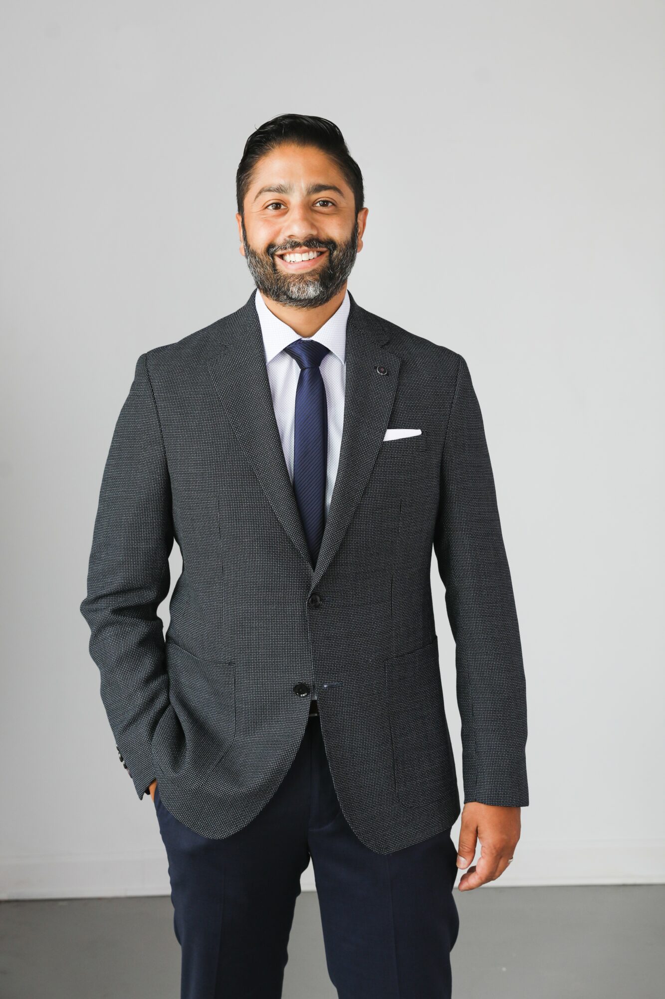 Man in a dark suit, white shirt, and tie smiling, standing against a plain light gray background.