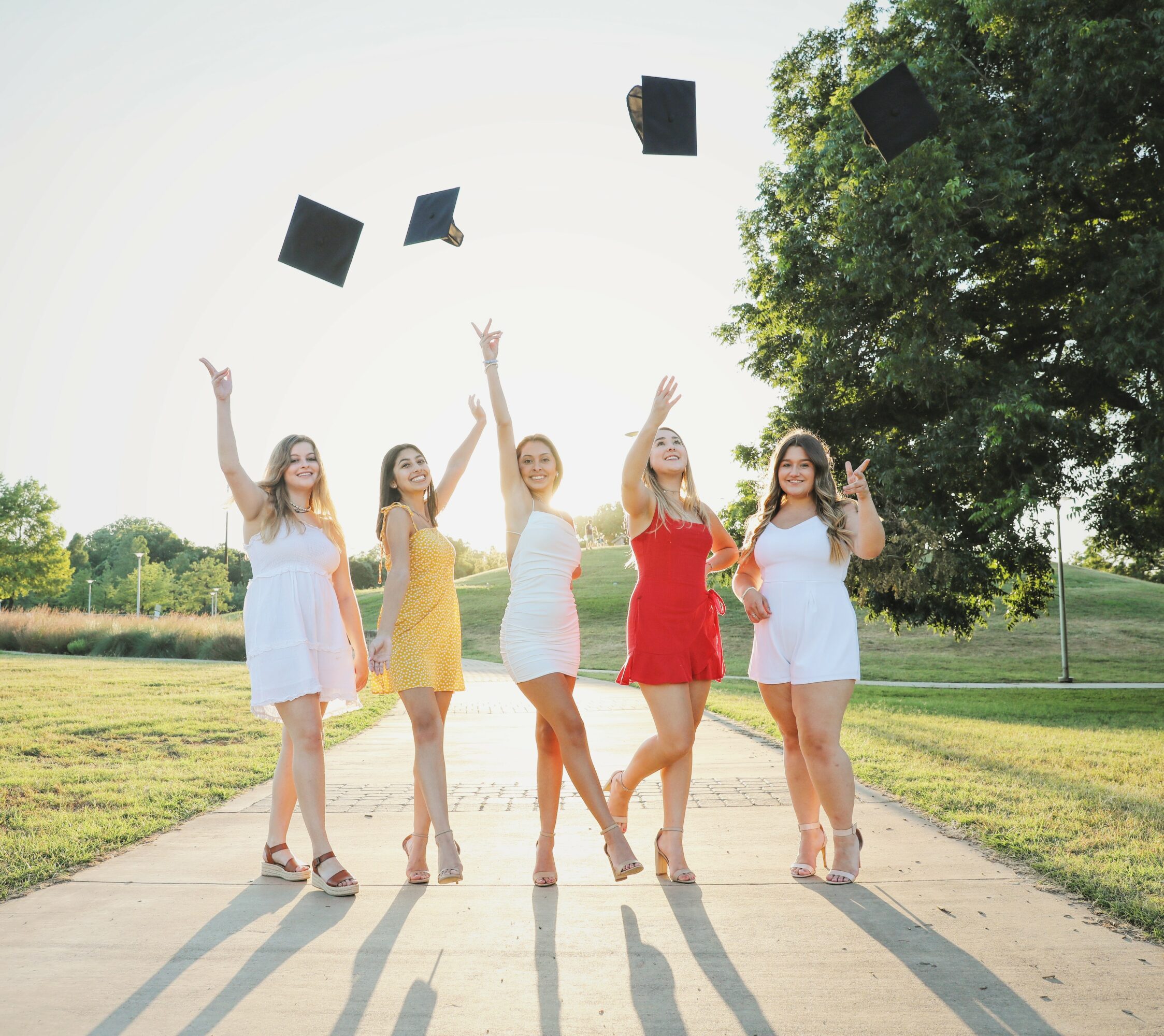 Five women in dresses celebrating graduation outdoors, tossing caps into the air on a sunny day, with trees and grass in background.