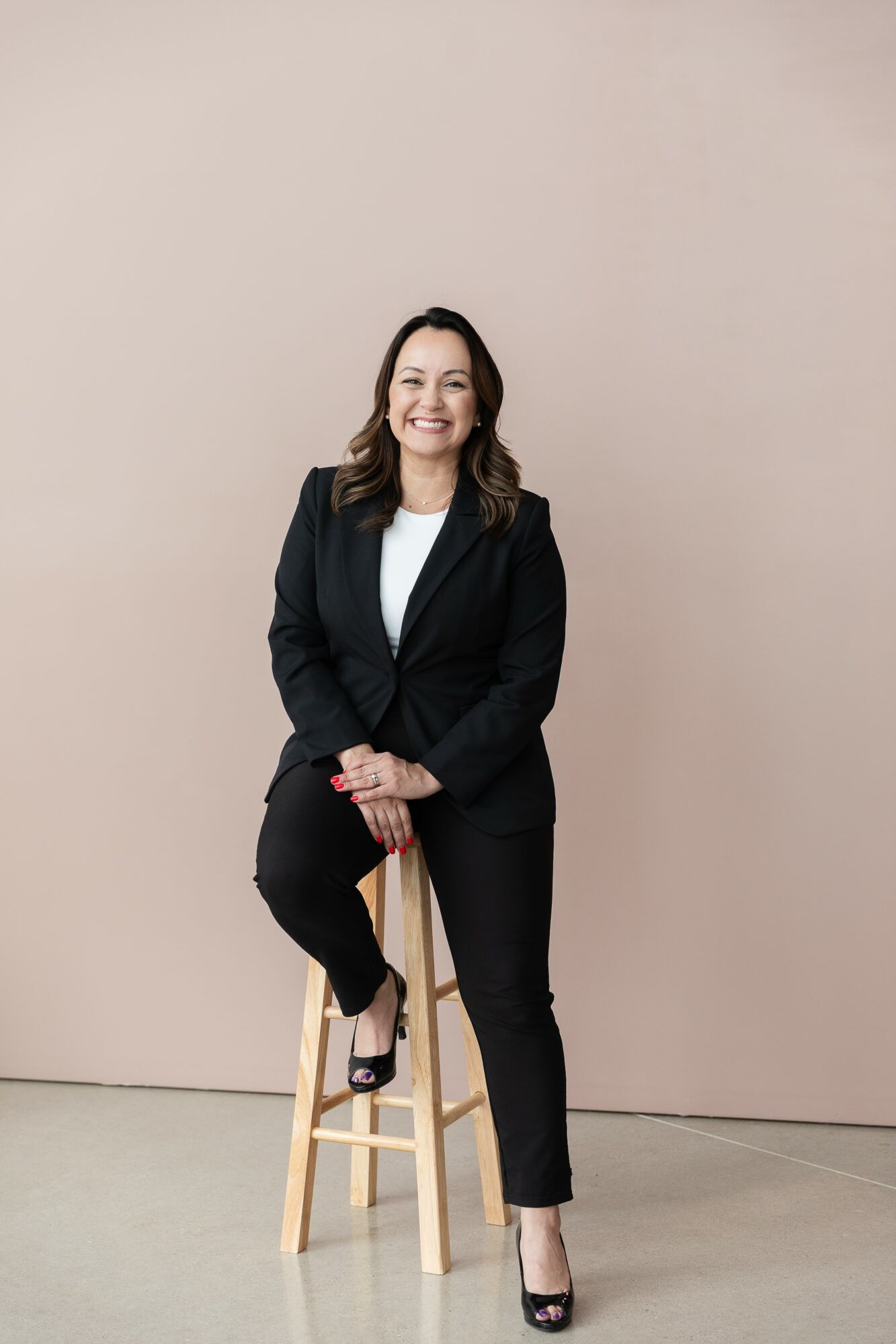 Woman in black suit sitting on a wooden stool, smiling, against a plain light background.