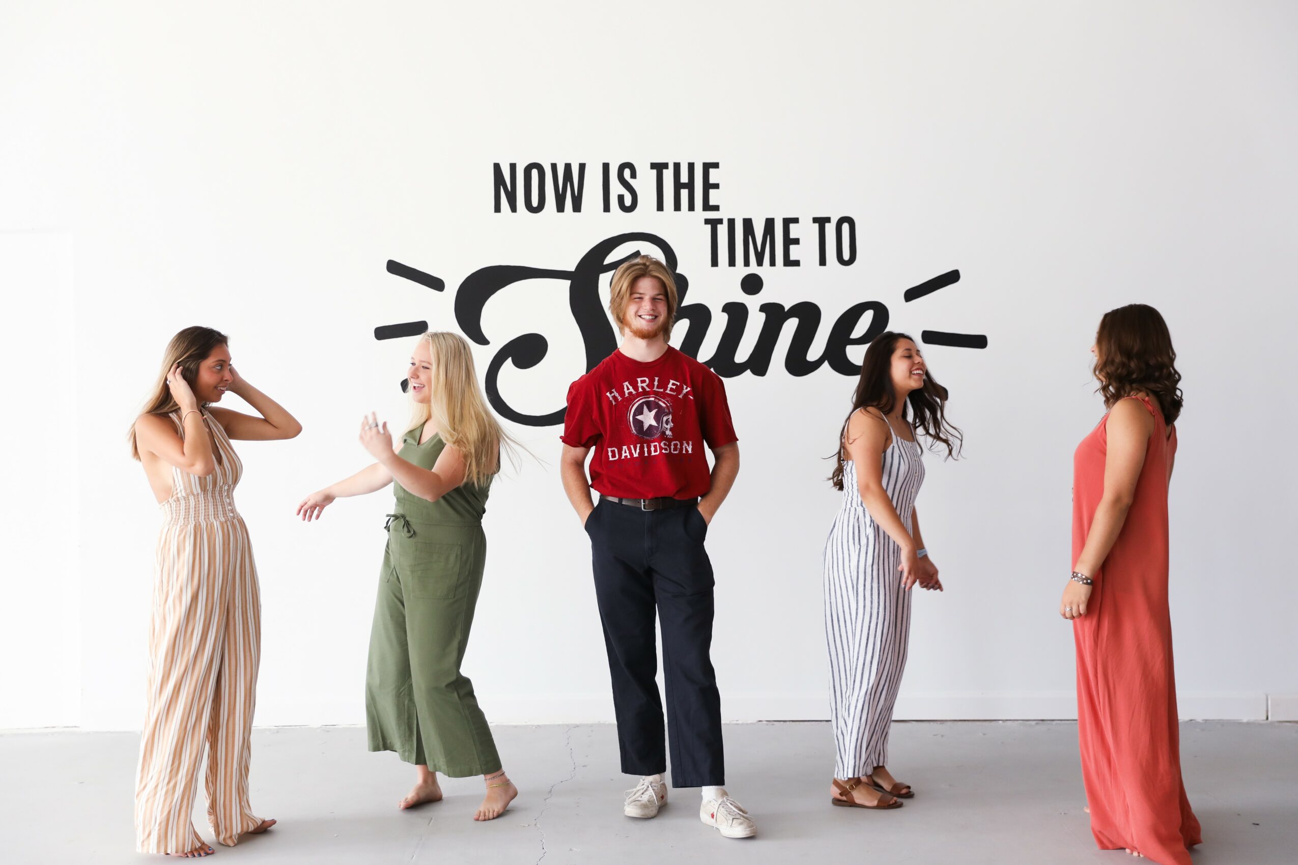 Five women stand in front of a white wall with black text reading 'Now is the time to online.' one woman wears a red shirt, others wear dresses, and they are engaged in conversation.