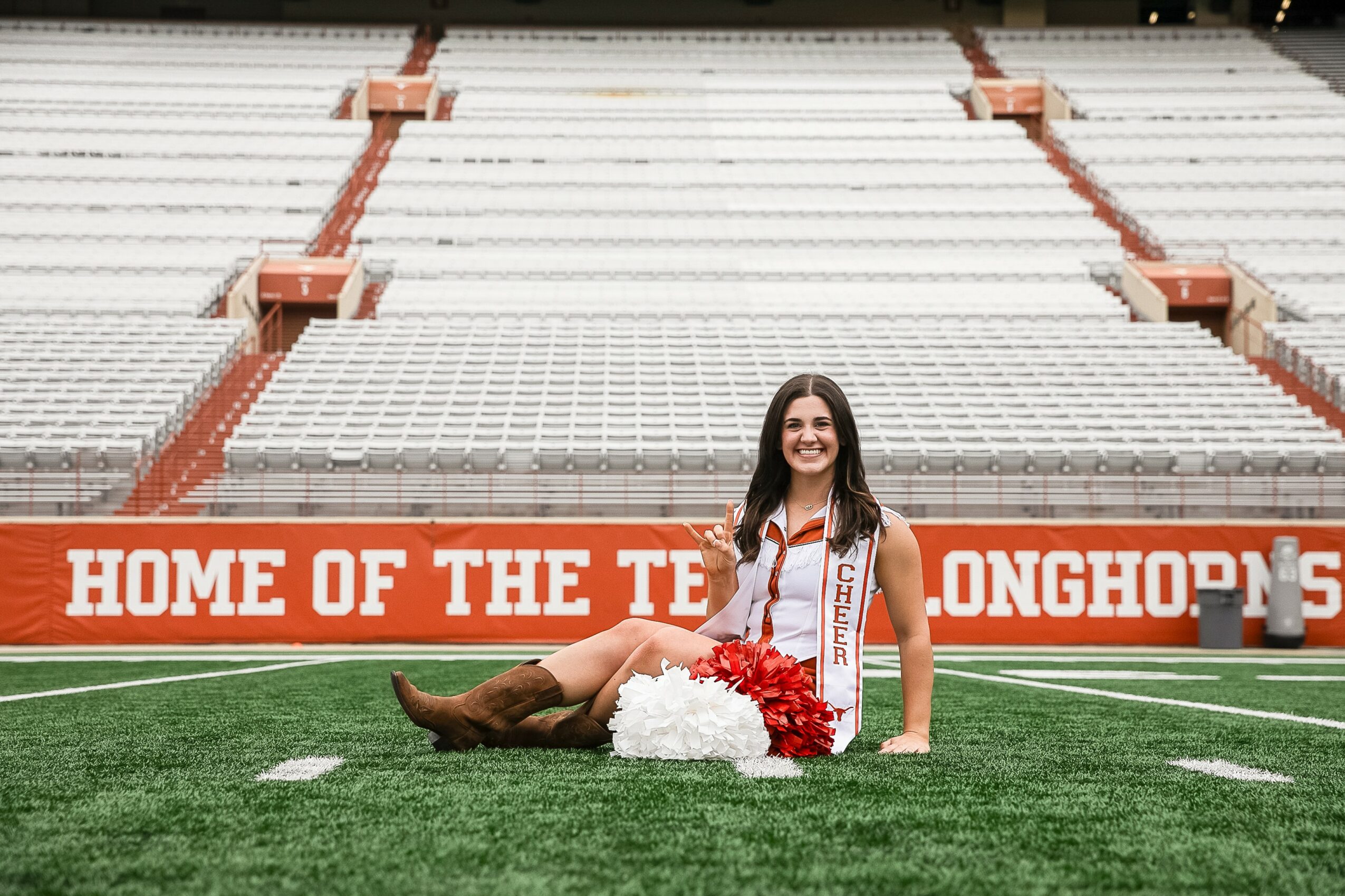 Young woman in cheerleading uniform sitting on football field with pom-poms, stadium seats in background, smiling.