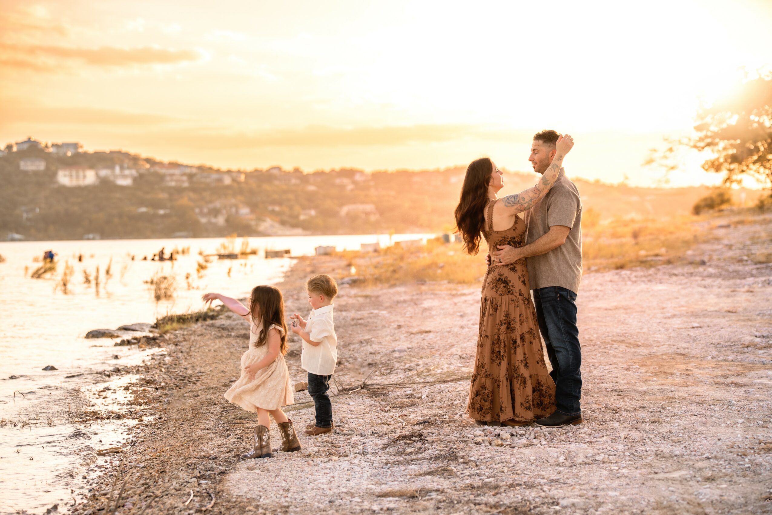 A family of four stands on a beach at sunset, with a cityscape in the background, children playing near the water.