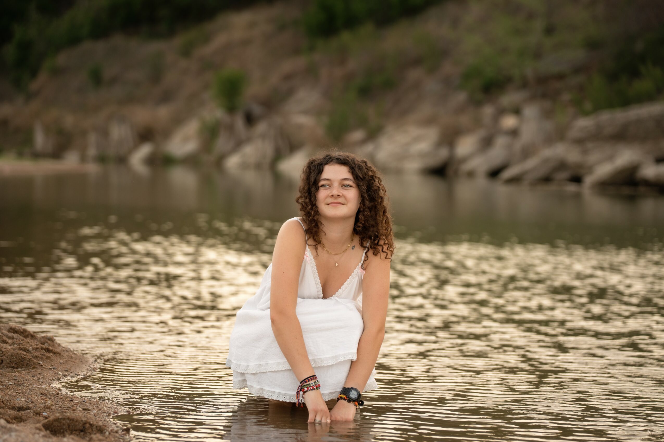Young woman with curly hair in a white dress kneeling in a river, smiling, with a rocky and green hillside background.
