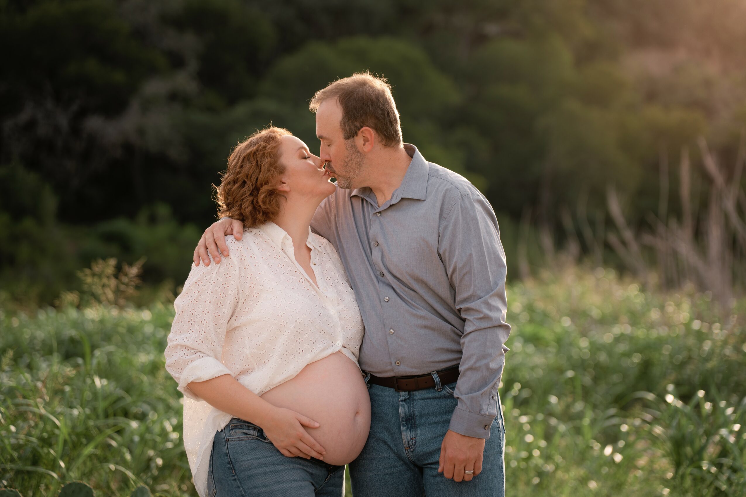 Pregnant woman and man kissing outdoors with greenery in background.