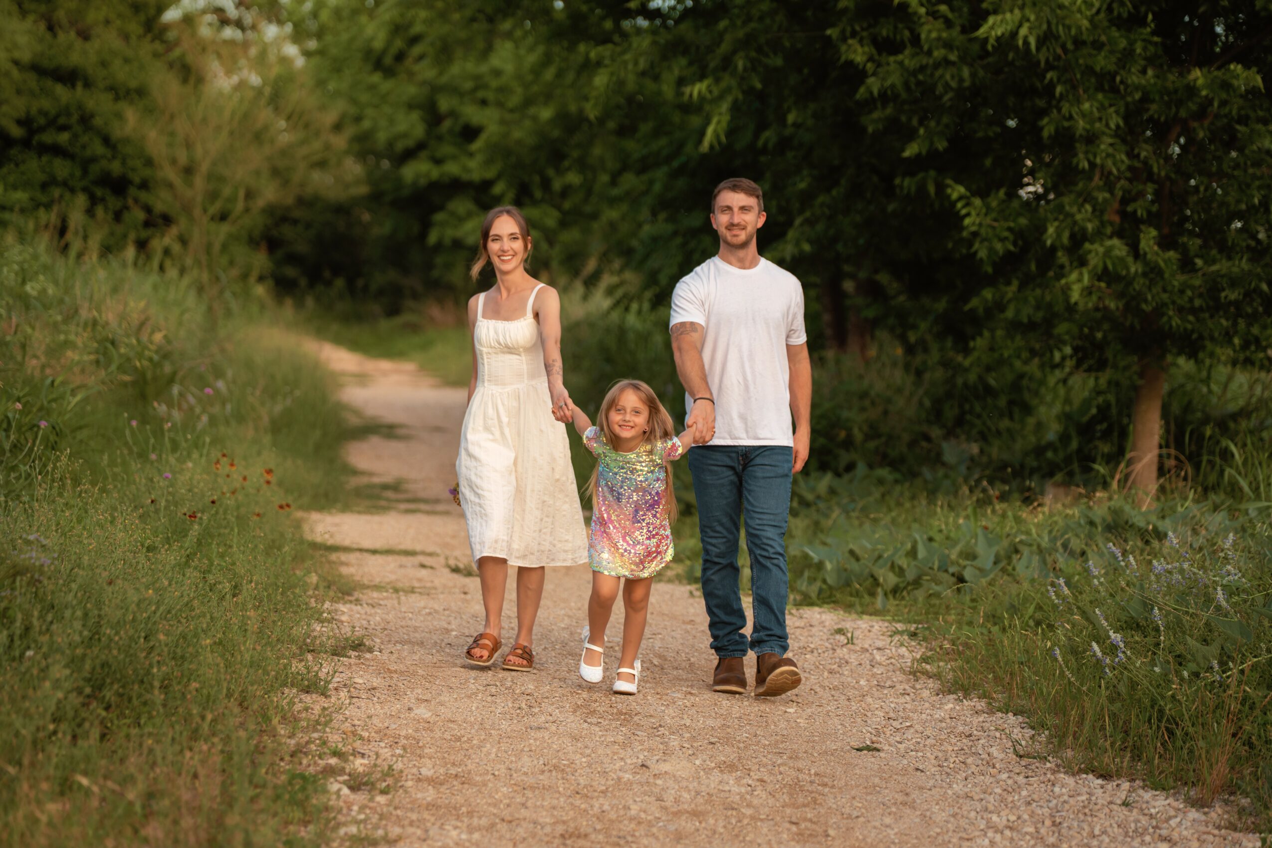 Family of three walking on a dirt path in a green outdoor setting, smiling, with trees and grass around.