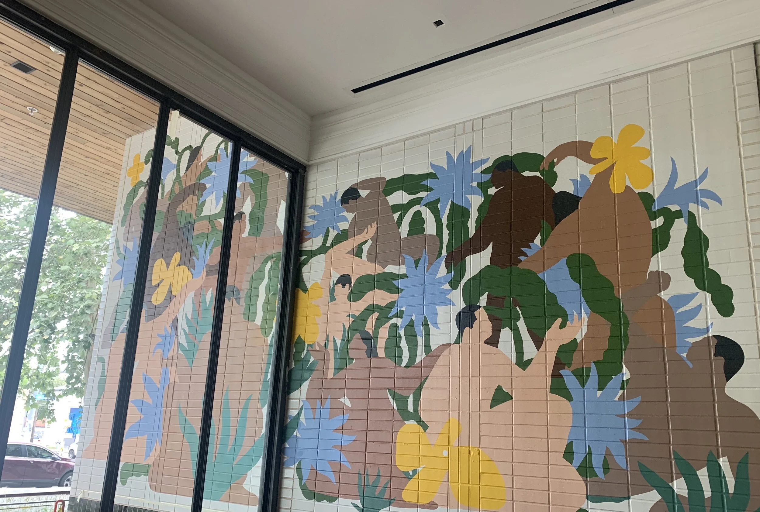 Colorful mural of abstract human figures and tropical plants on a wall near a window.