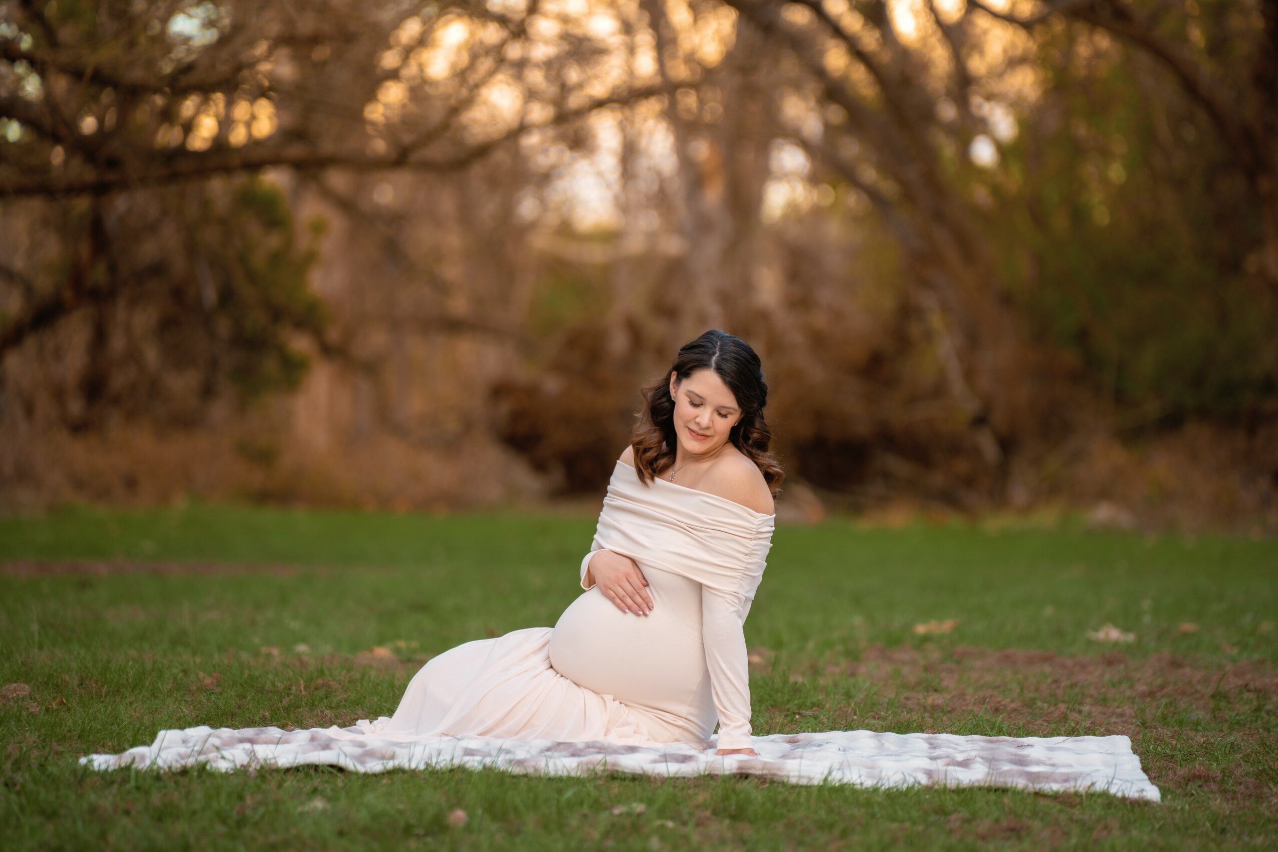 Pregnant woman in a white dress sitting on a blanket outdoors with trees in the background.