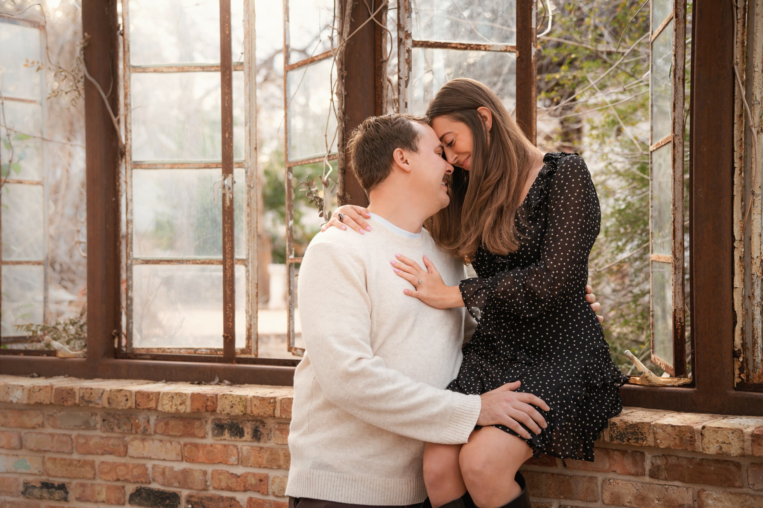 Two women sit close together on a brick windowsill, smiling and touching foreheads in a cozy indoor setting.