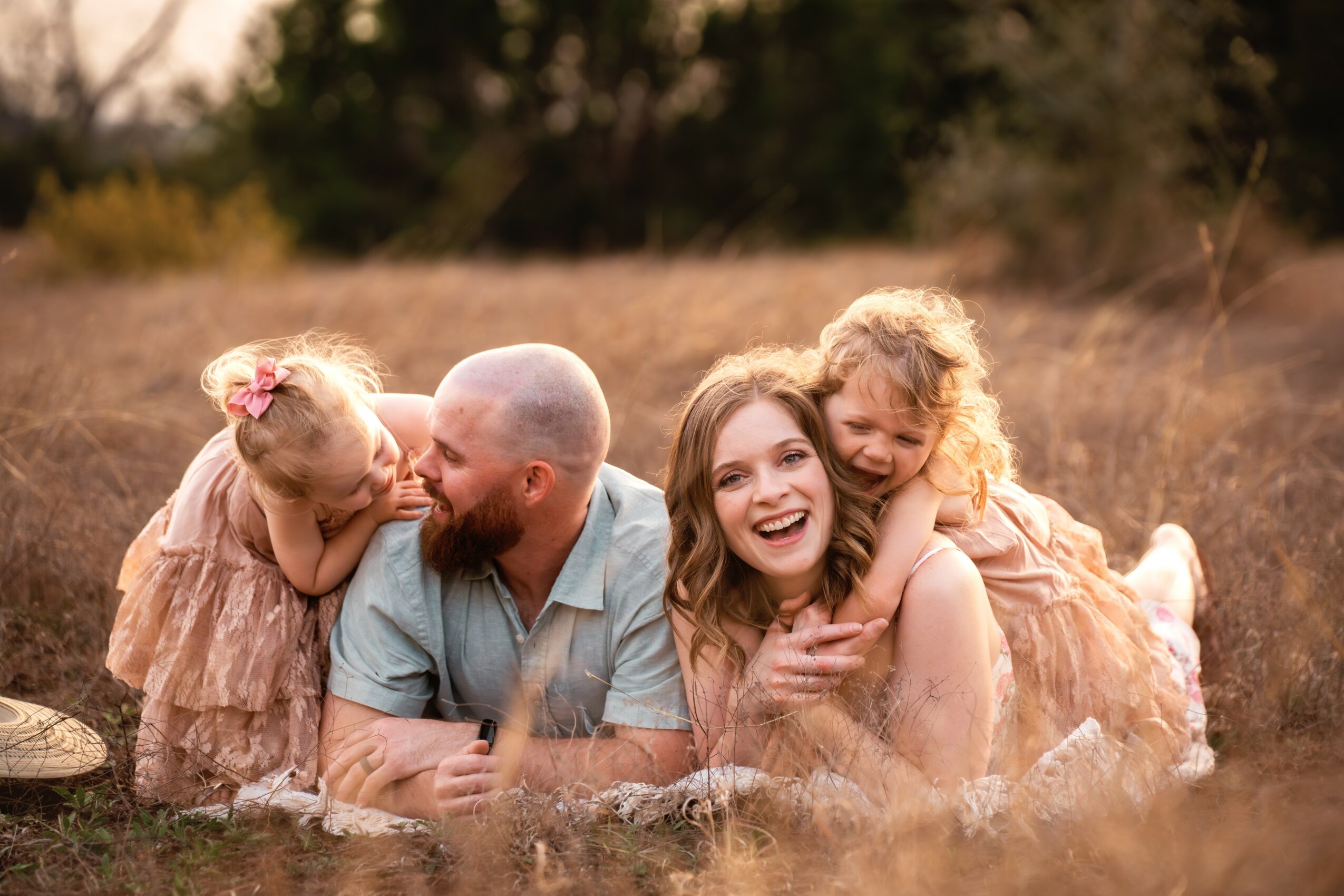 A family of four lying on the grass outdoors, smiling and hugging each other, with trees in the background.
