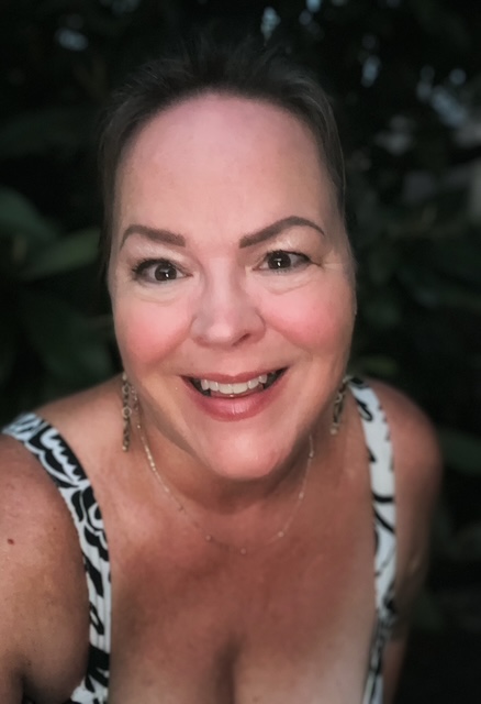 Smiling woman with dark hair, wearing a patterned top, outdoors with dark foliage background.
