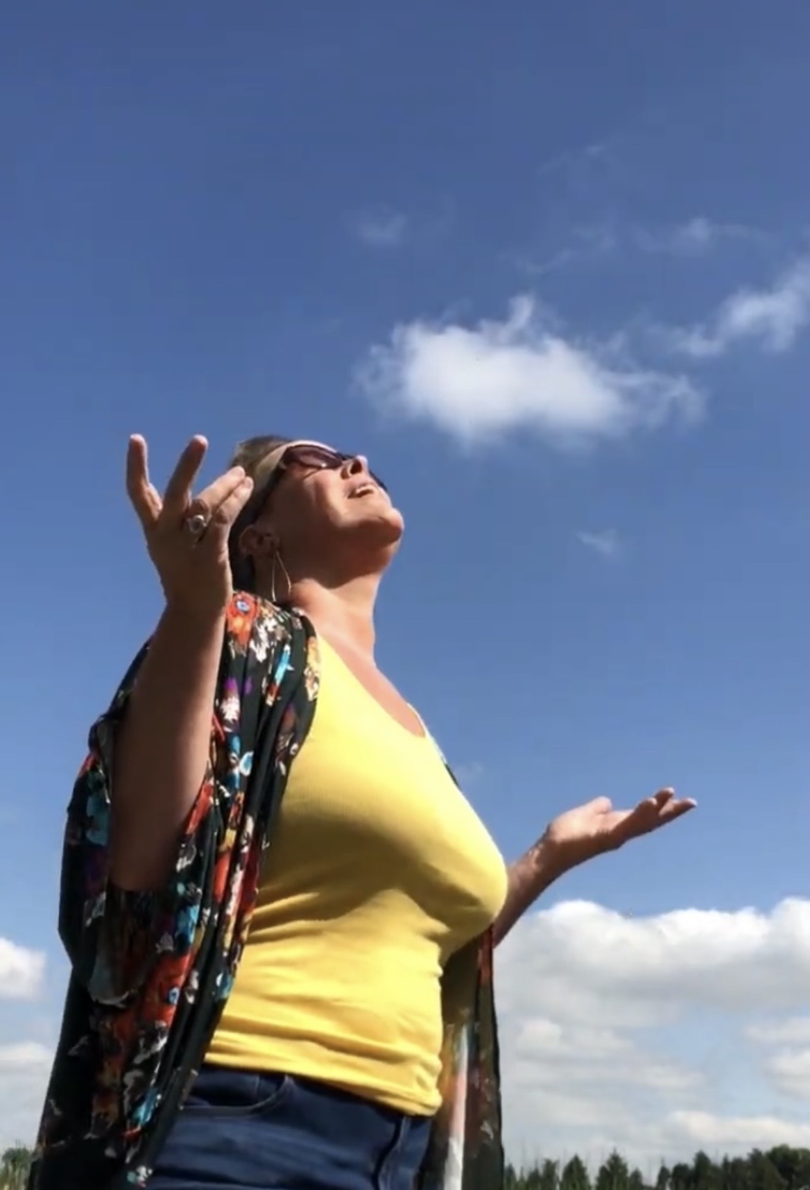 Woman outdoors with arms raised, face tilted upward, wearing a yellow top and a colorful shawl, blue sky background.