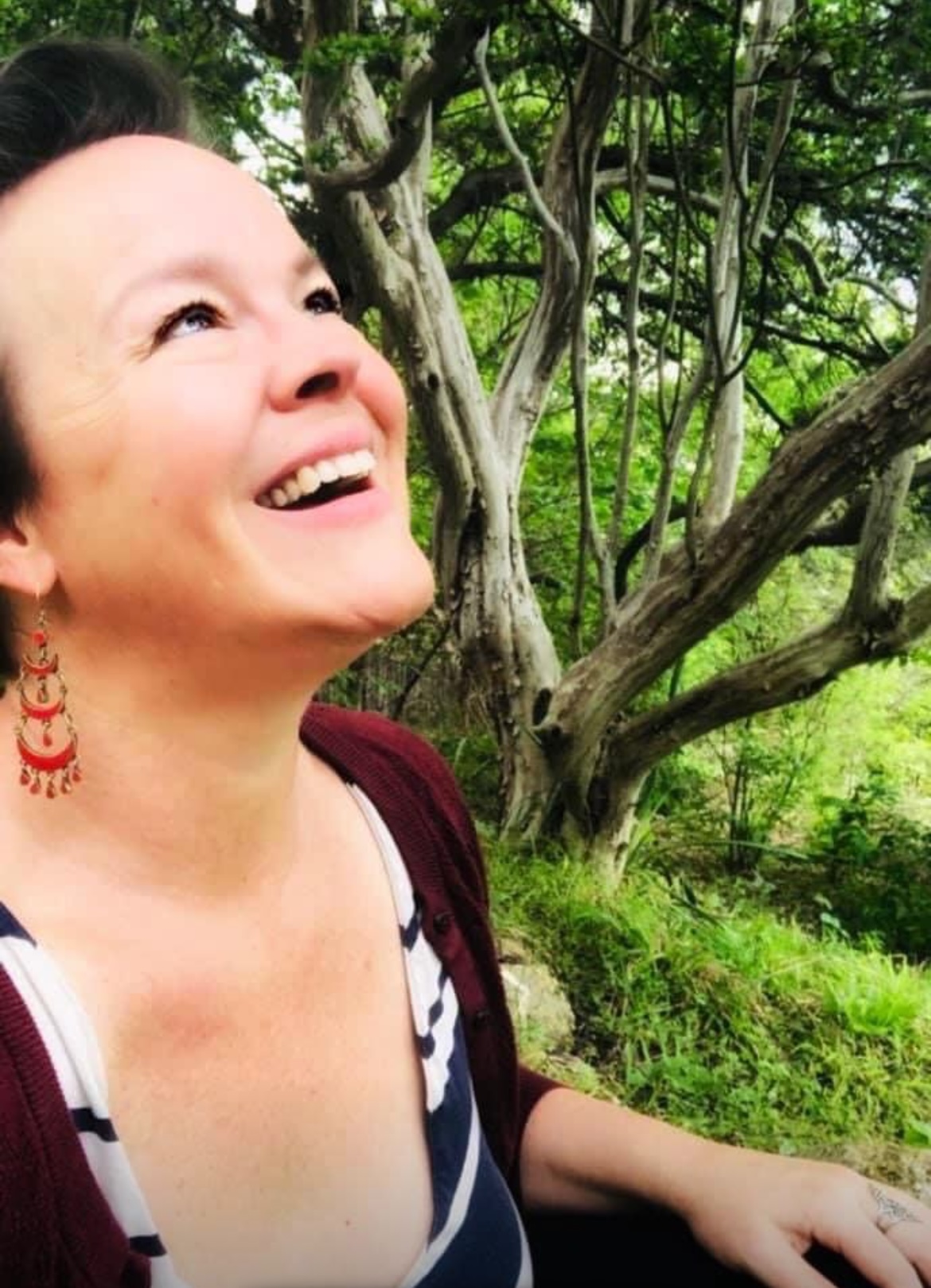Woman smiling outdoors with trees and greenery in the background.