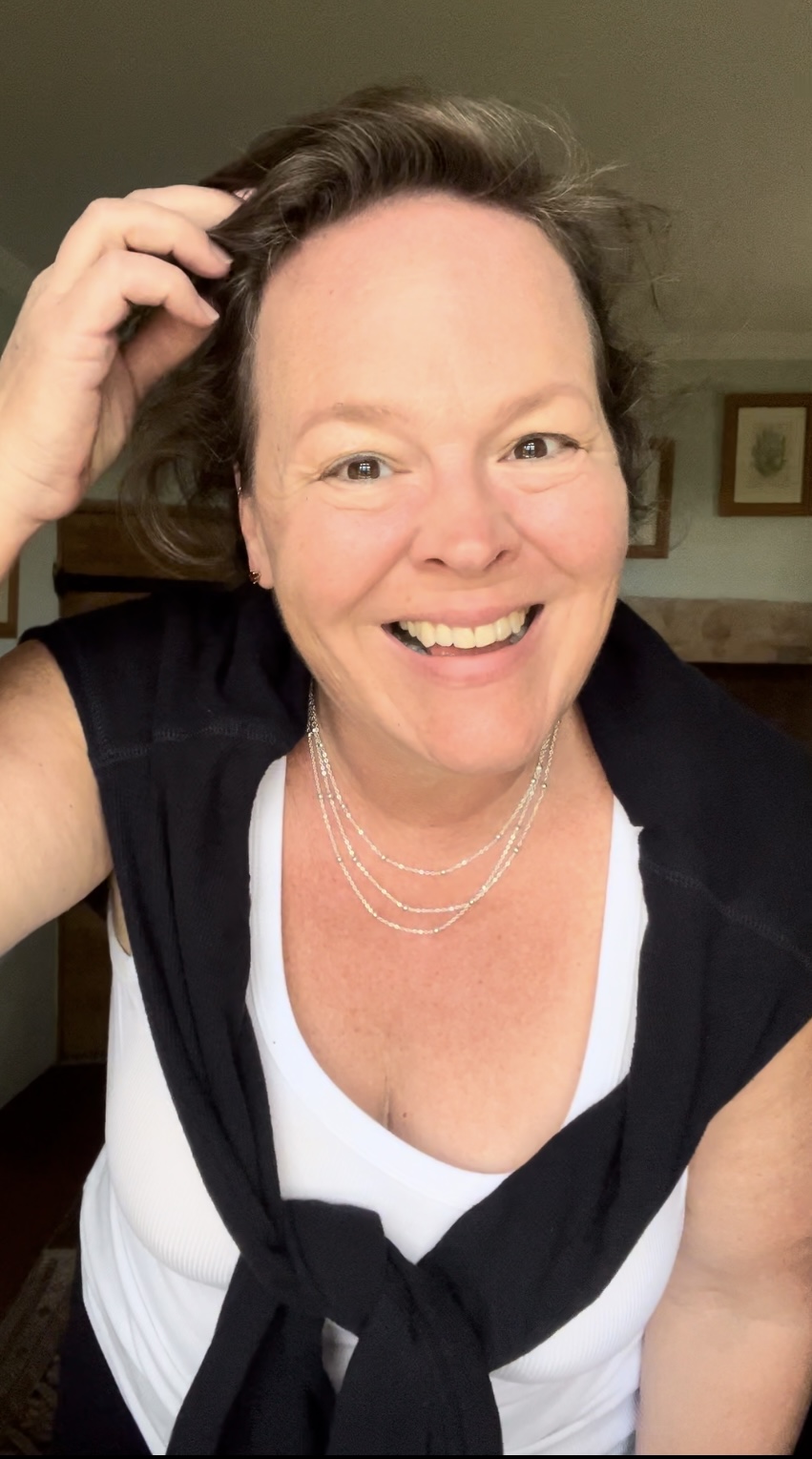 Smiling woman with short dark hair, wearing a white top and layered necklaces, touching her hair.