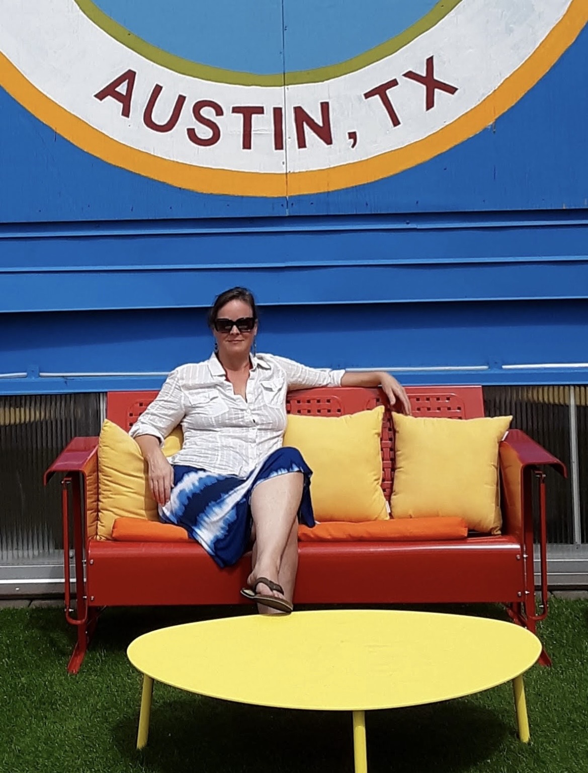Woman sitting on a red and yellow outdoor sofa with yellow cushions, in front of a blue wall with a sign reading 'AUSTIN, TX'.