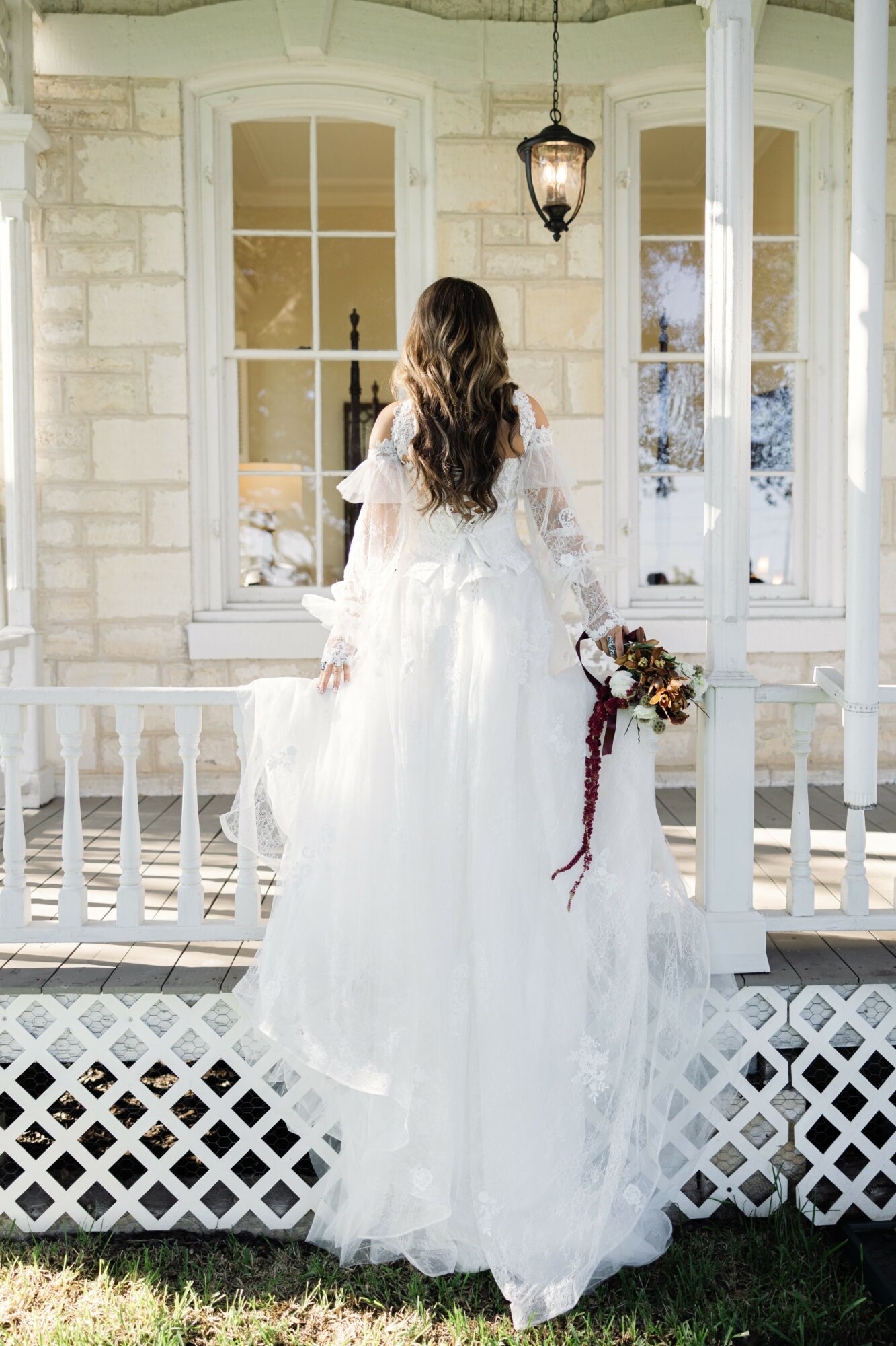 Woman in a white dress with long hair standing on a porch, holding a bouquet, facing away from the camera.