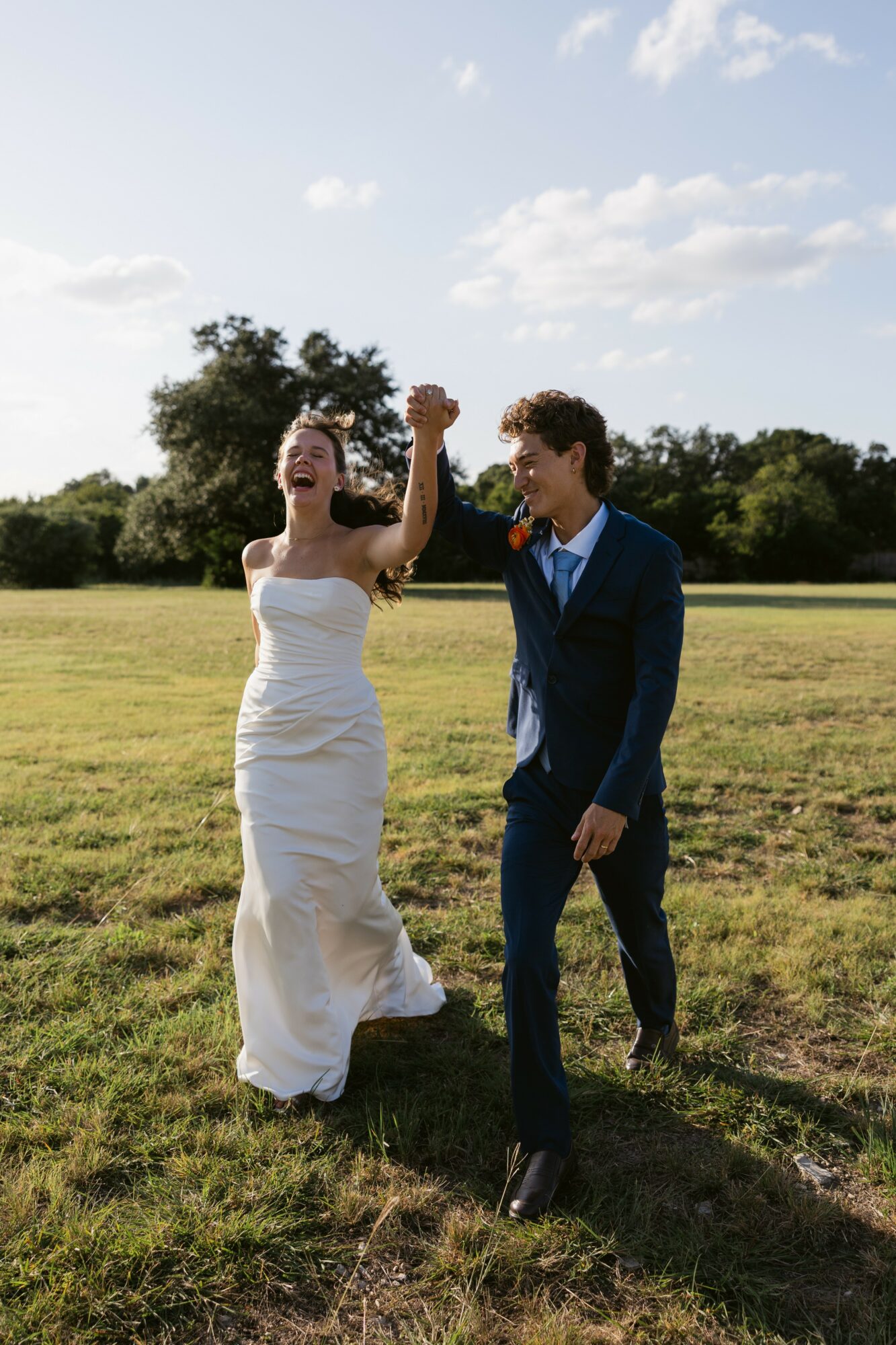 A woman in a white dress and a man in a blue suit are celebrating outdoors, holding hands and smiling.