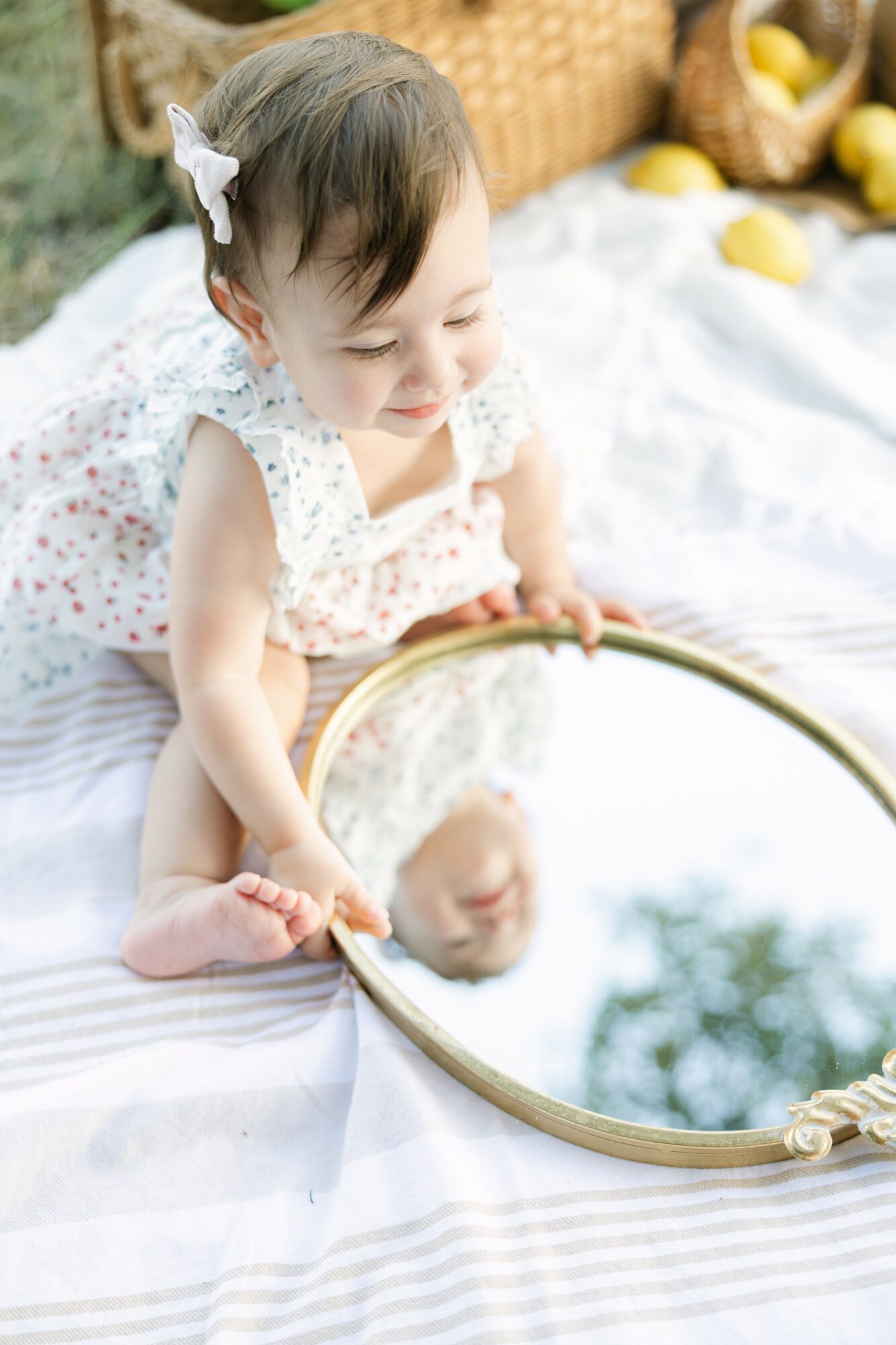 Young girl with a bow in her hair looking into a mirror, outdoors on a white blanket, with yellow fruits nearby.