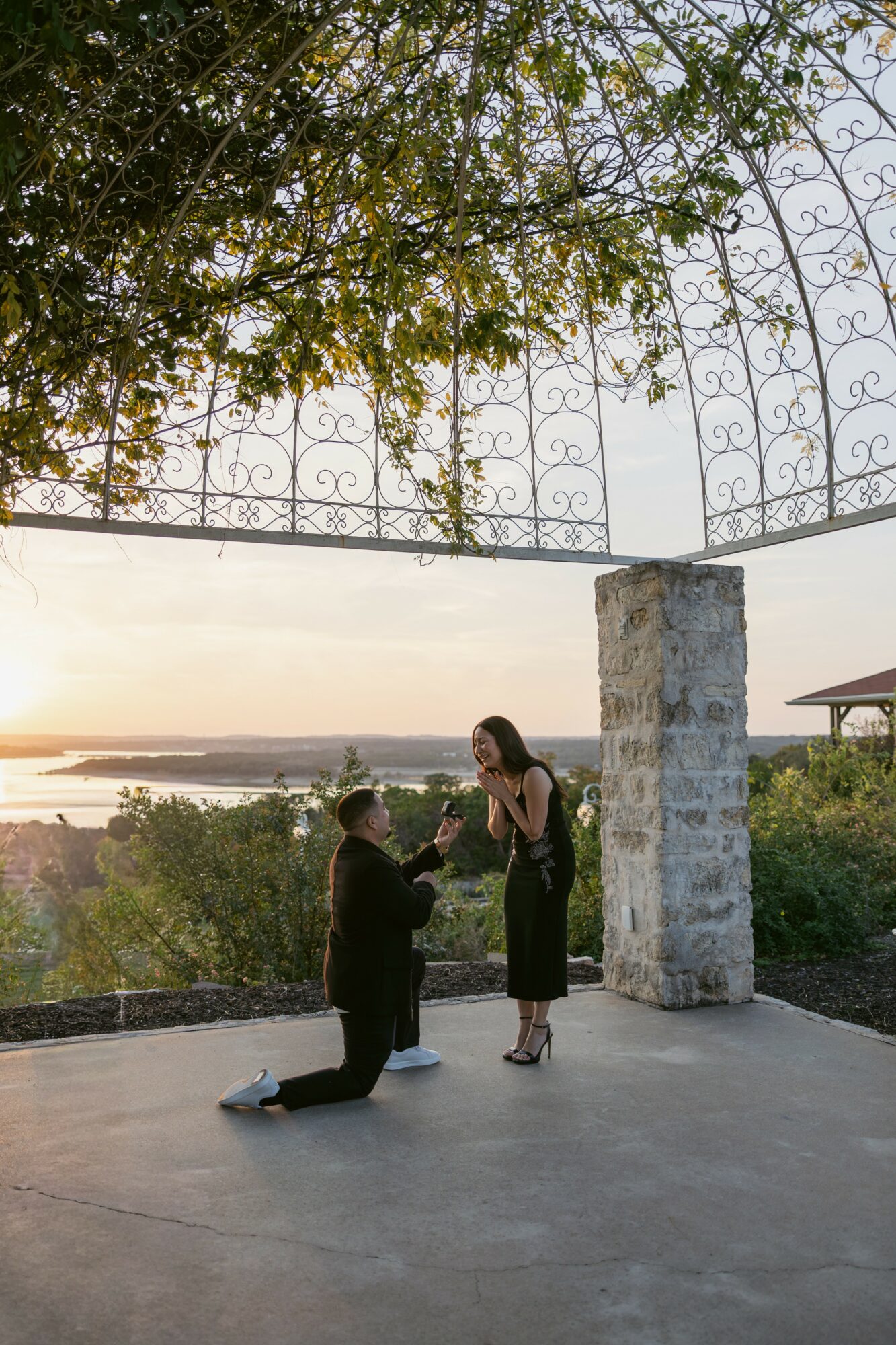 A person kneeling and proposing to a woman outdoors during sunset, with a scenic landscape in the background.