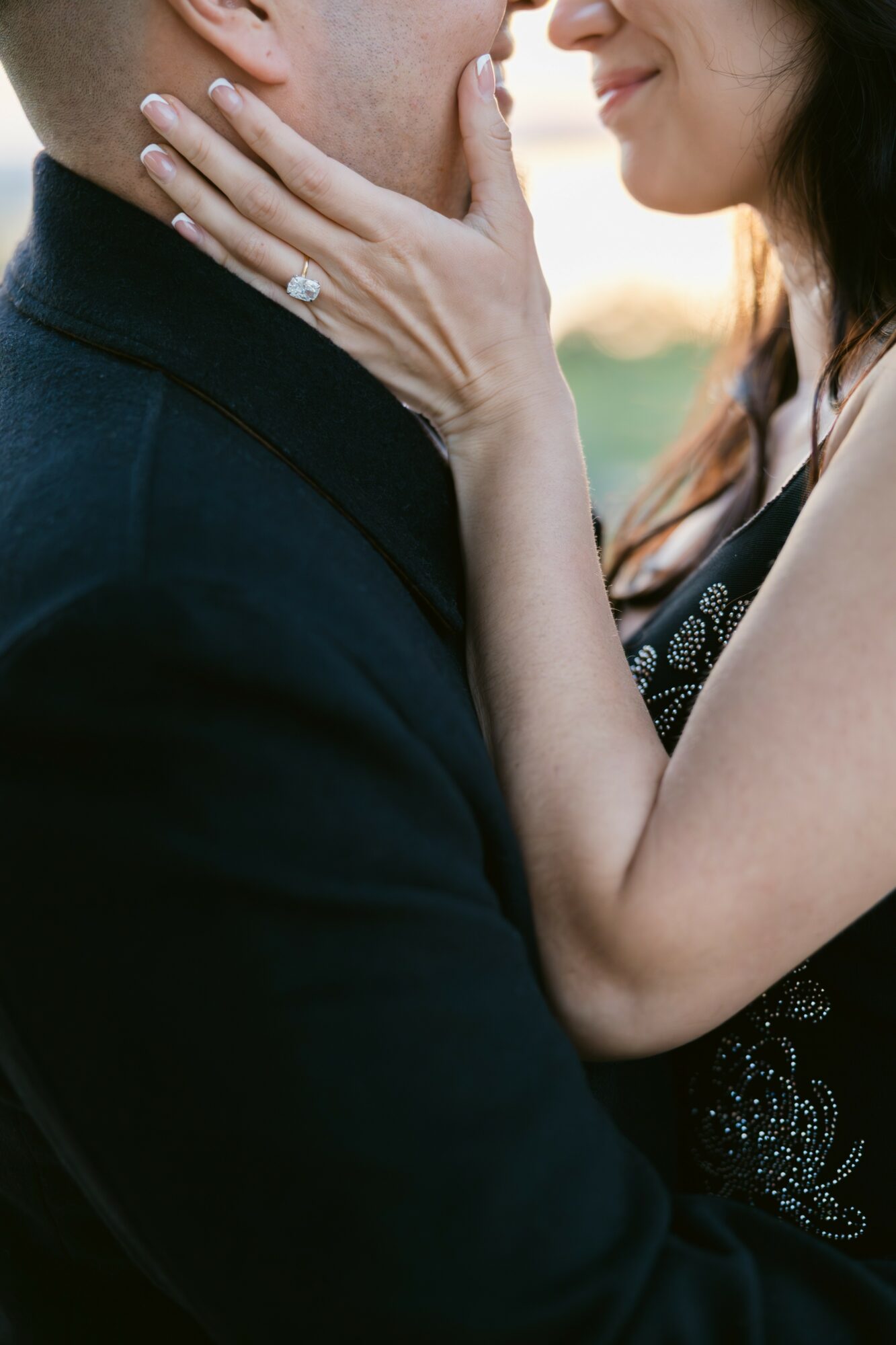 Close-up of a woman gently touching a man's face, both smiling softly, outdoors during sunset, with a blurred background.
