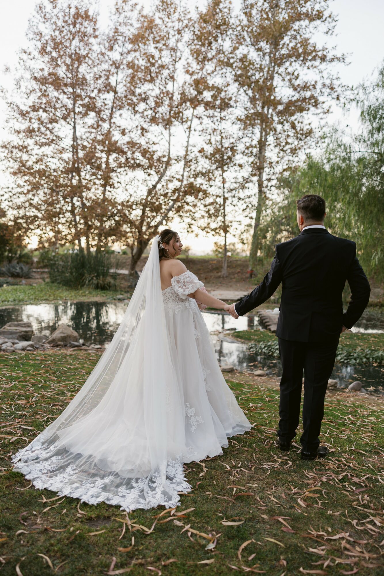 Bride and groom holding hands outdoors near a lake with trees and sunset in background.