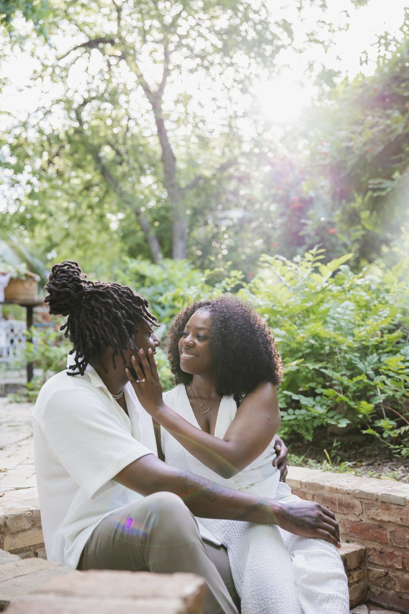 Two women sit outdoors surrounded by greenery, smiling and touching faces, sunlight filtering through trees, in a peaceful setting.