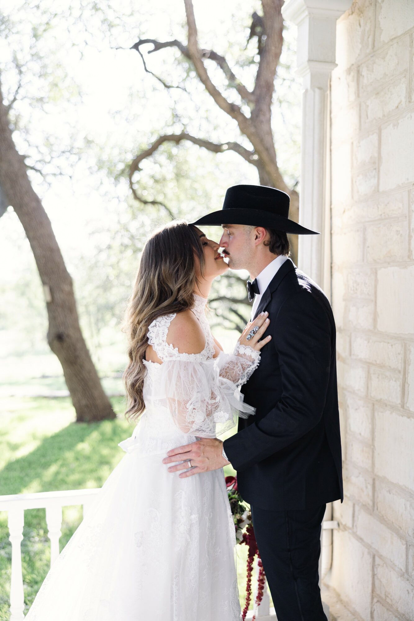 Two women in wedding attire standing close, facing each other, outdoors near a stone wall and trees.