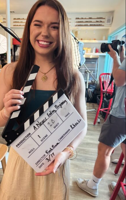 Young woman smiling holding a film clapperboard in a room with colorful chairs and a person in the background.