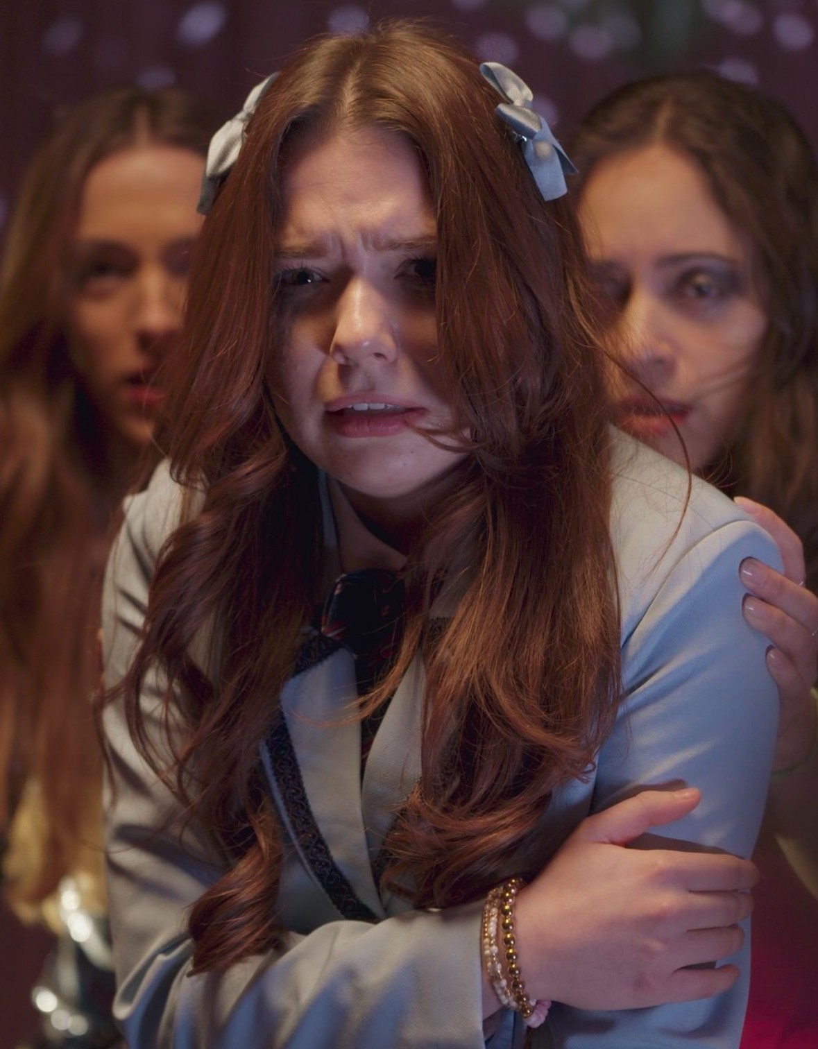 Three young women, one in the foreground with long brown hair and a distressed expression, two others behind her, in a dimly lit setting.