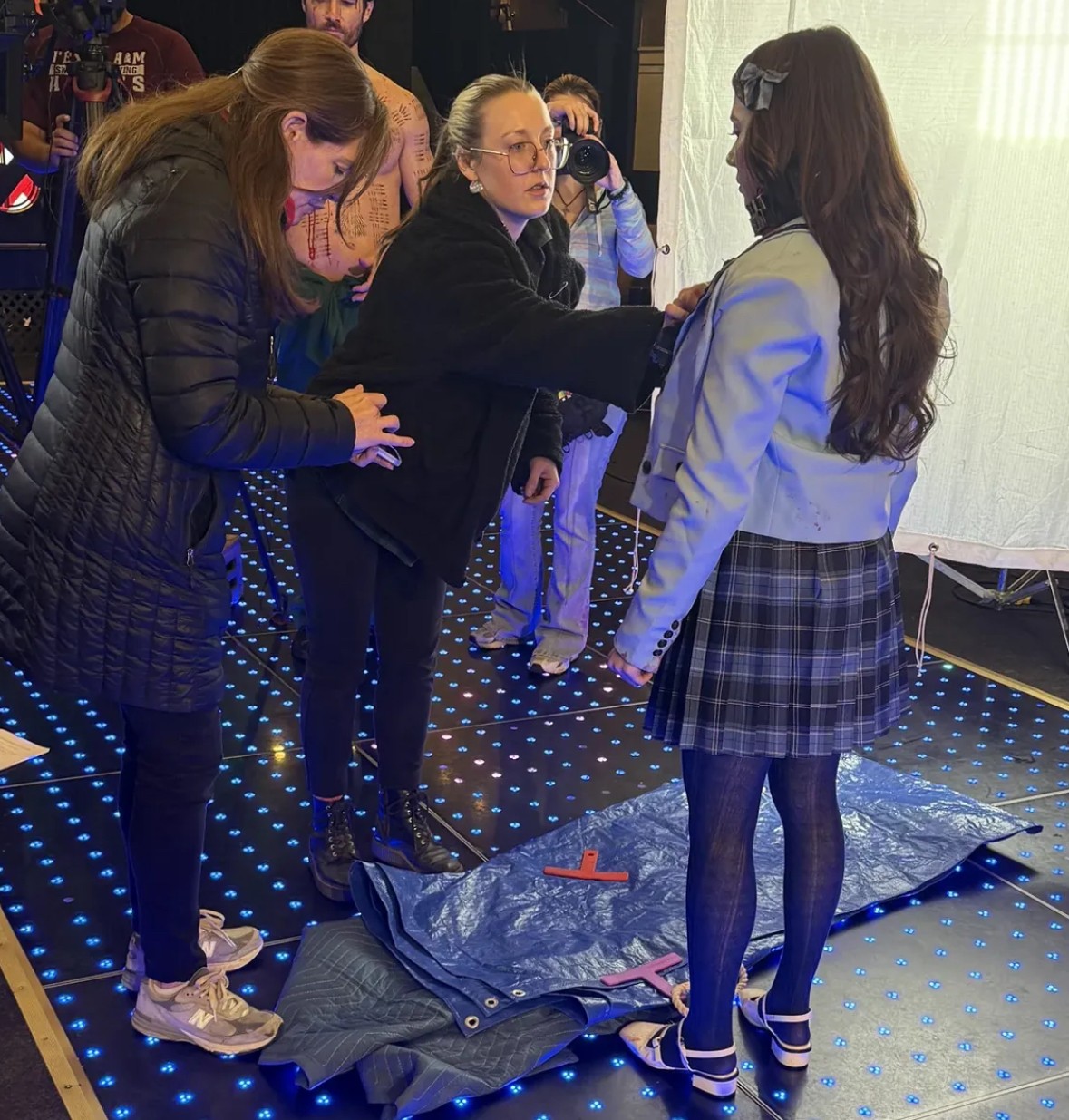 Three women standing around a blue mat with a red cross, indoors with a dotted floor, engaged in conversation.