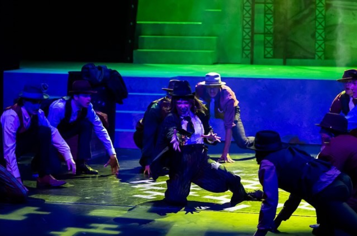 Group of people on stage with green and blue lighting, some kneeling and others standing, in a performance setting.