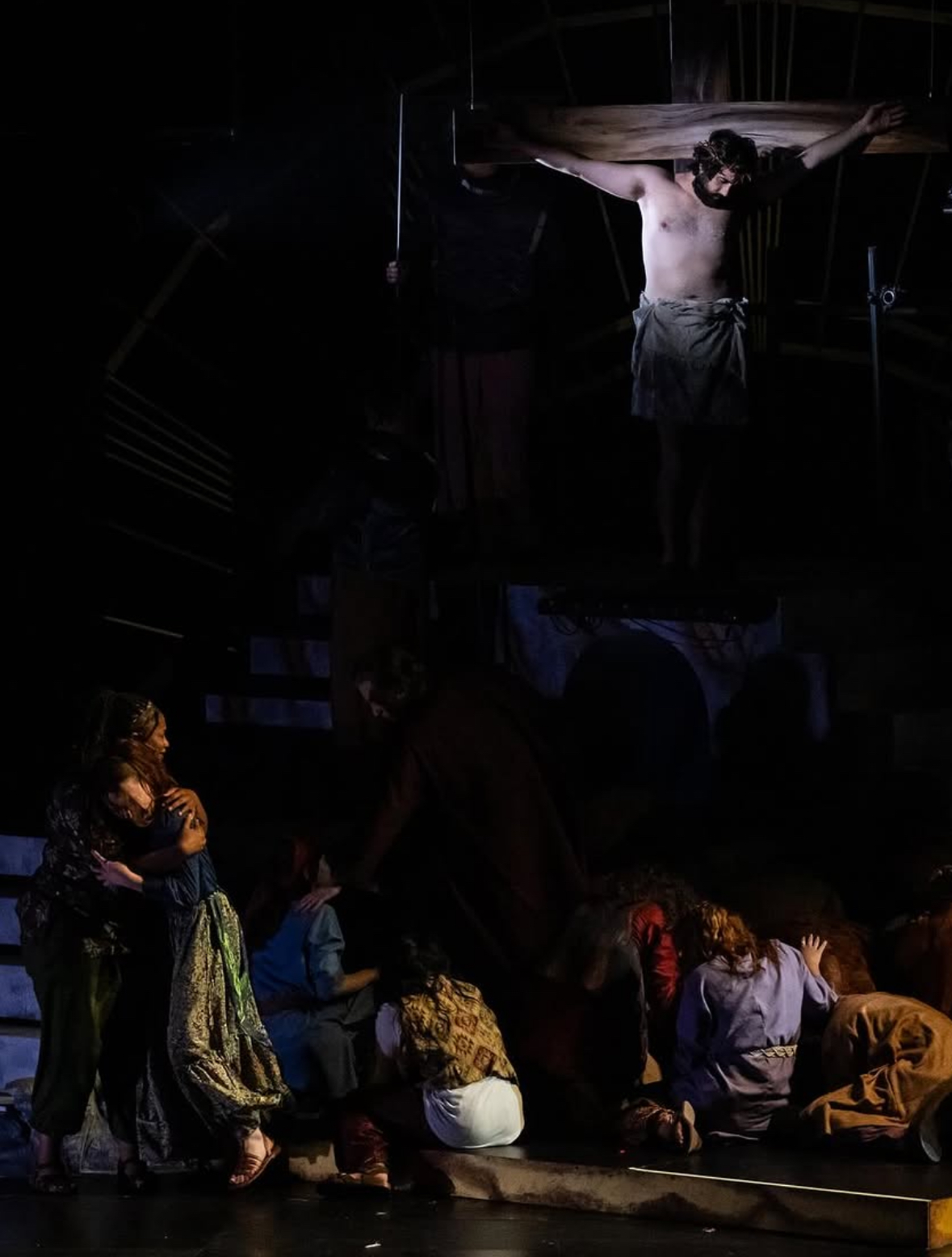 Stage scene with a person on cross and group of people sitting and standing nearby, dim lighting, theatrical setting.