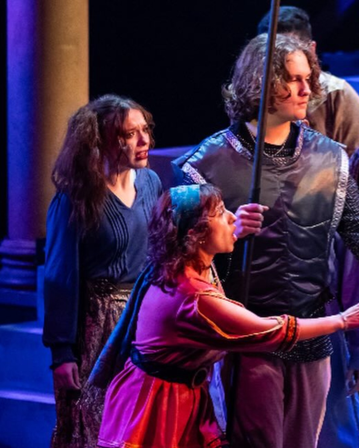 Three people on stage, two women and a girl, with dark hair, in a theatrical setting with blue and purple lighting.
