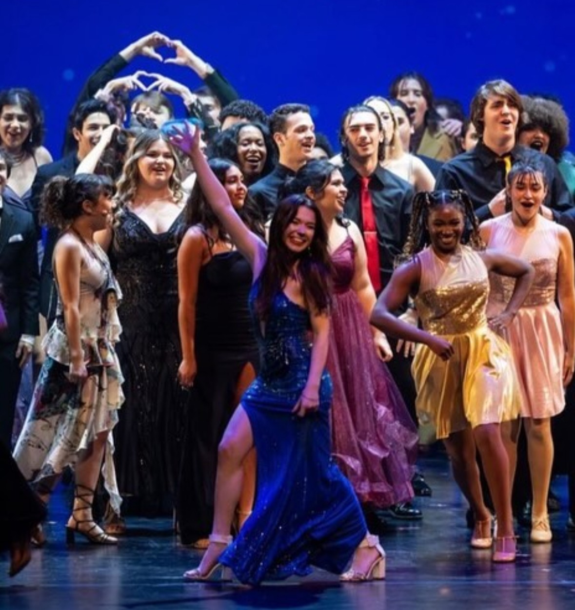 Group of diverse people dancing on stage, smiling, with a blue background, some forming heart shapes with their hands.