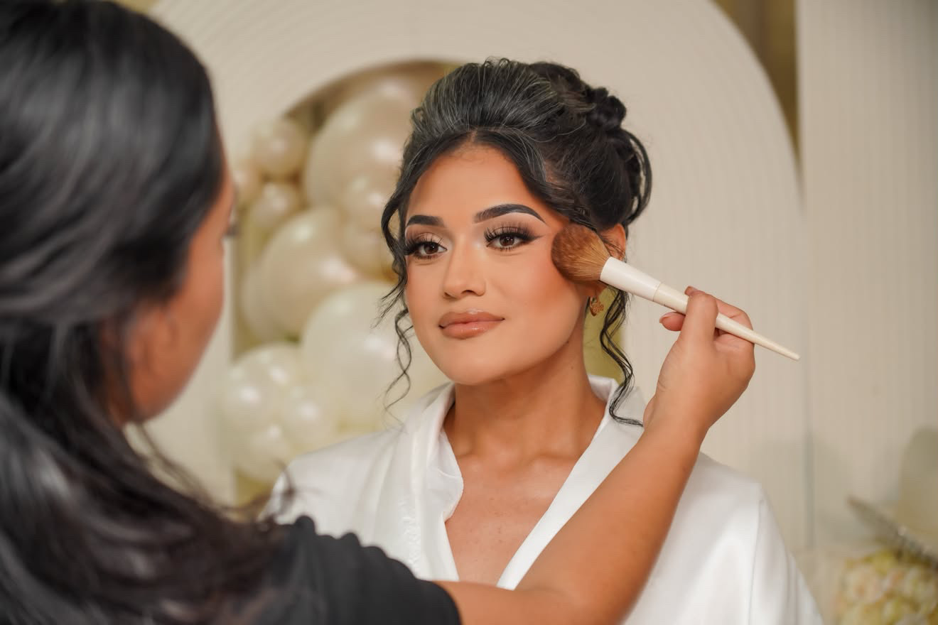 A woman with dark hair and makeup is having her face gently touched with a brush by another person, in a well-lit room.