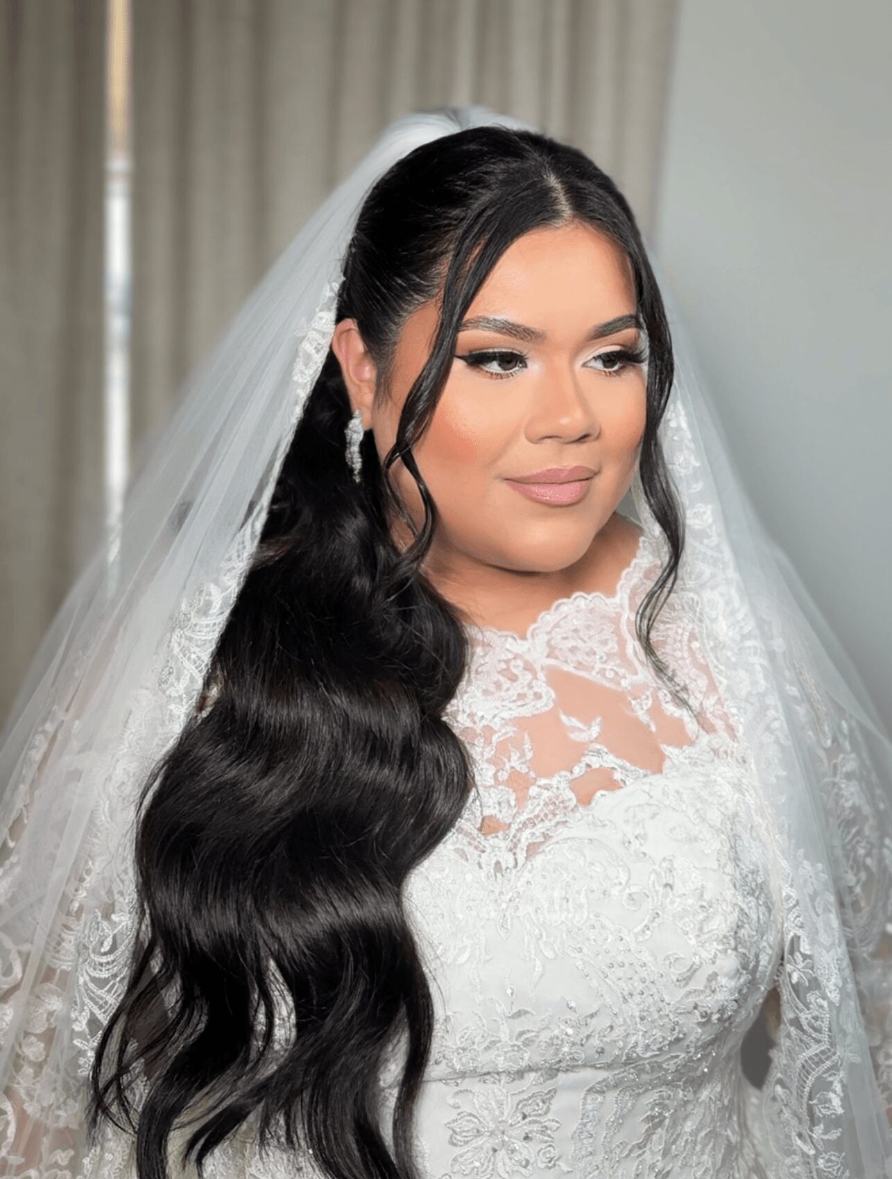 Bride with long, wavy dark hair, wearing a lace wedding dress and veil, standing indoors with curtains in background.