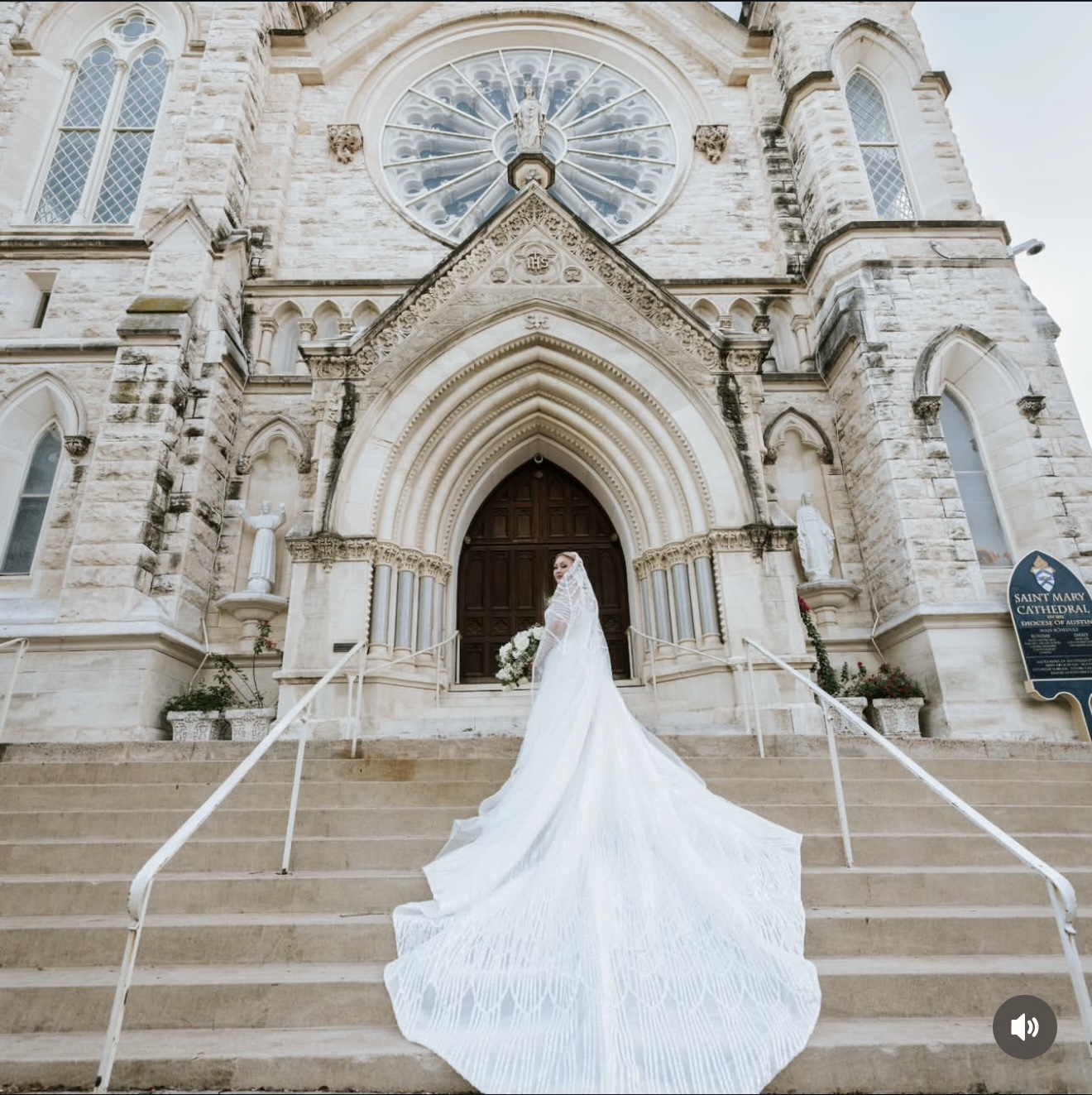 Bride in a white wedding gown with a long train stands on church steps, facing the entrance with ornate architecture.