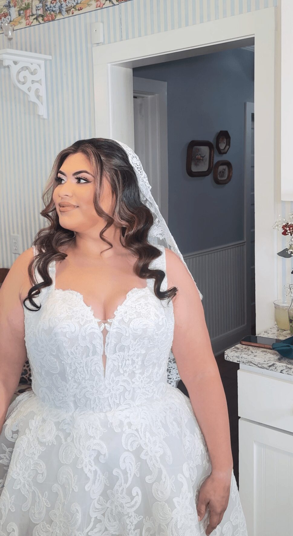 Bride in wedding dress with dark hair and veil, standing indoors near a doorway and a white cabinet.