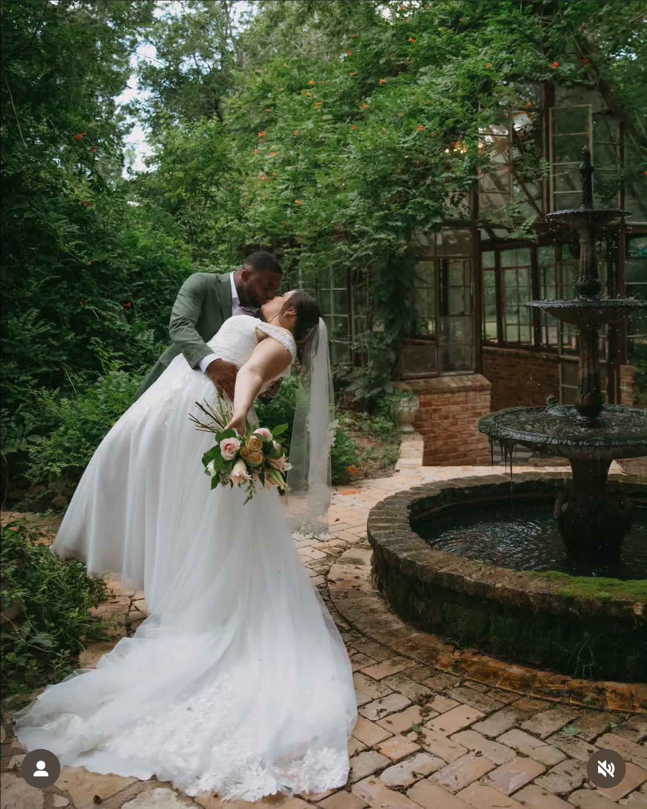 Bride and groom kiss outdoors near a fountain, surrounded by trees and greenery, with a brick pathway and garden structure.