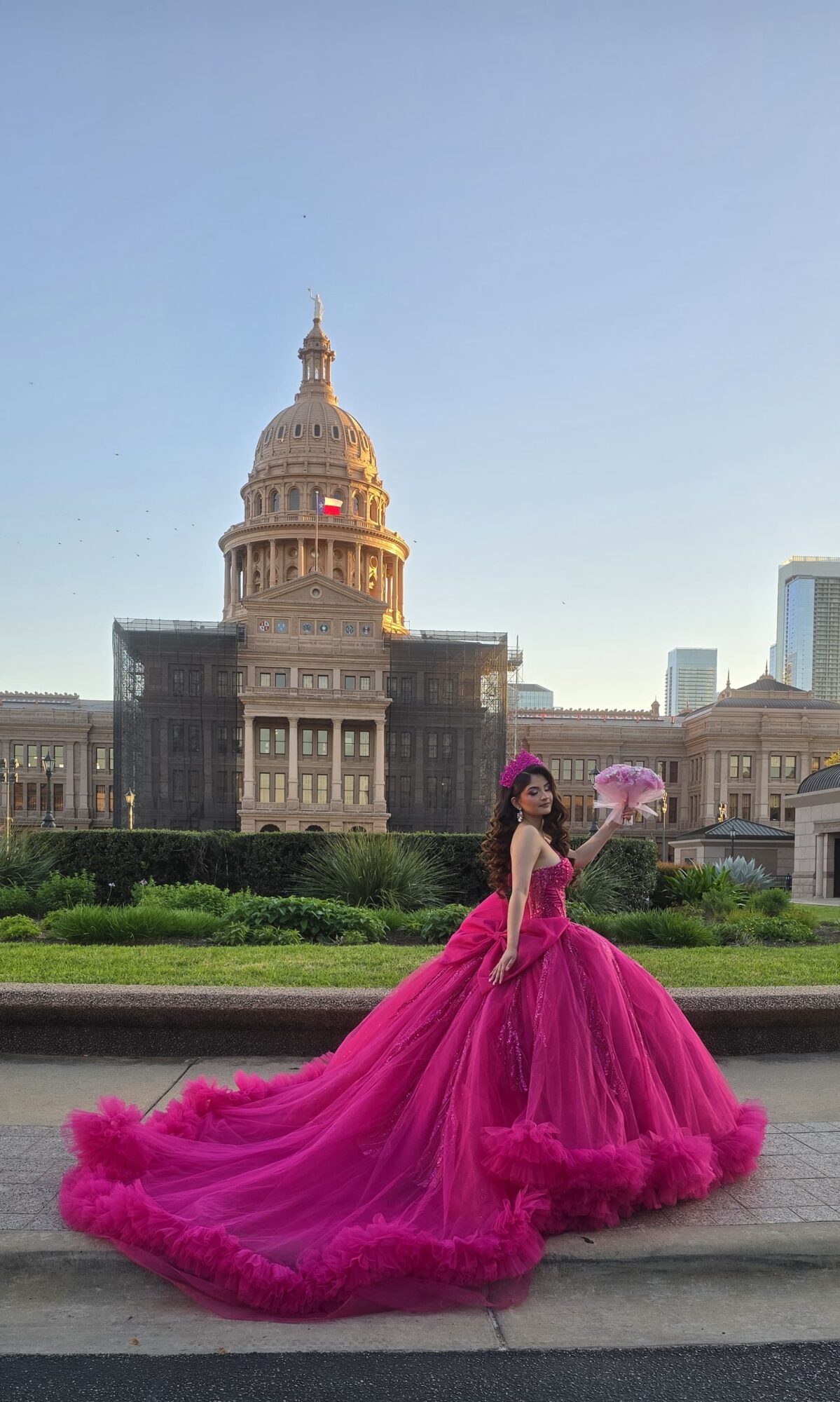 Woman in a pink gown holding a parasol in front of a historic building with a dome, greenery, and a clear sky.